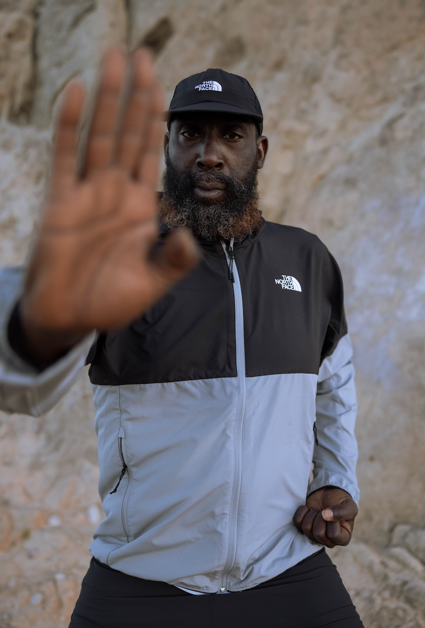 A man wearing a black and gray The North Face jacket and cap making a stop gesture with his left hand while looking directly at the camera, standing against a rocky background.