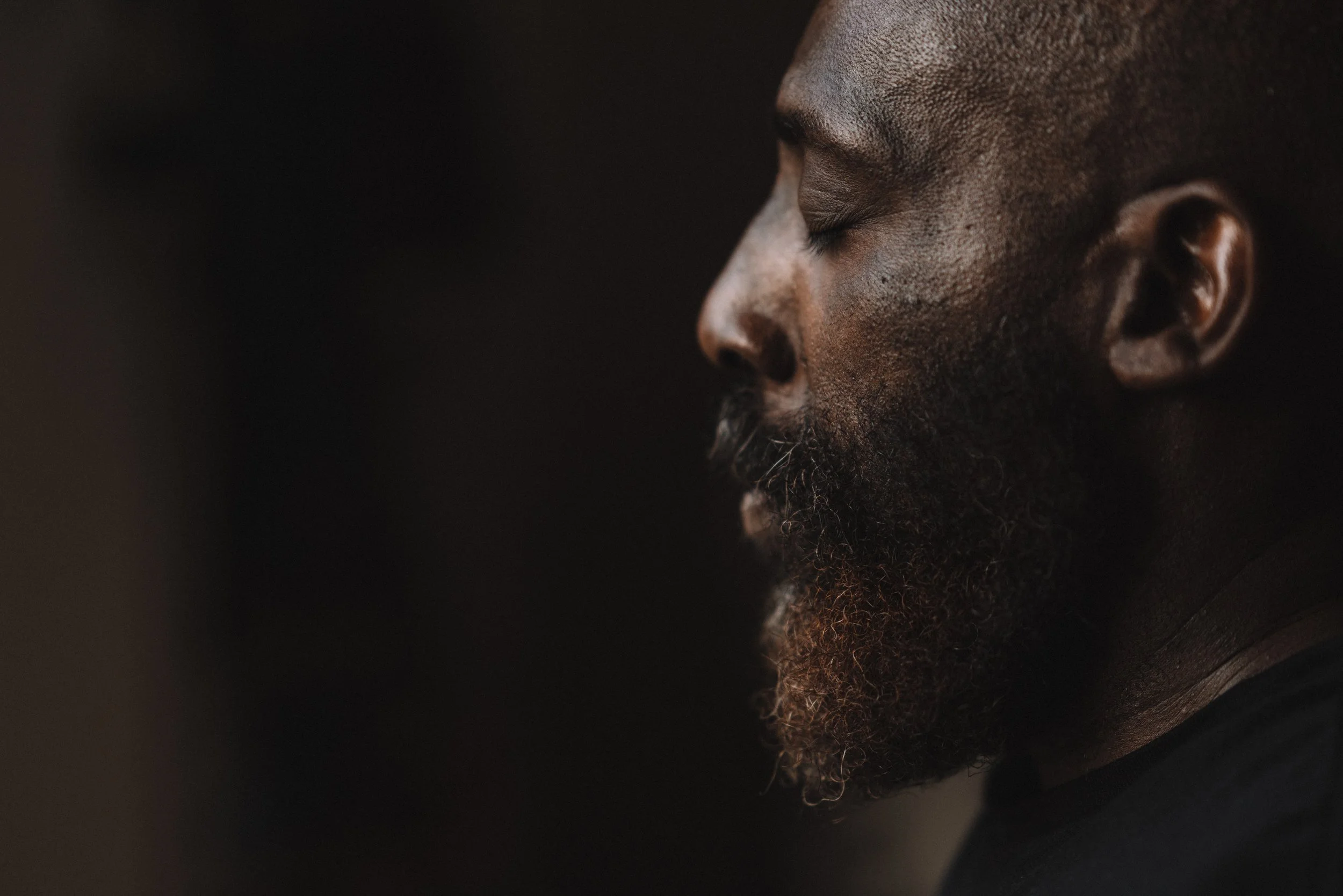 Close-up side profile of a man's face with dark skin and a beard, with his eyes closed against a dark background.