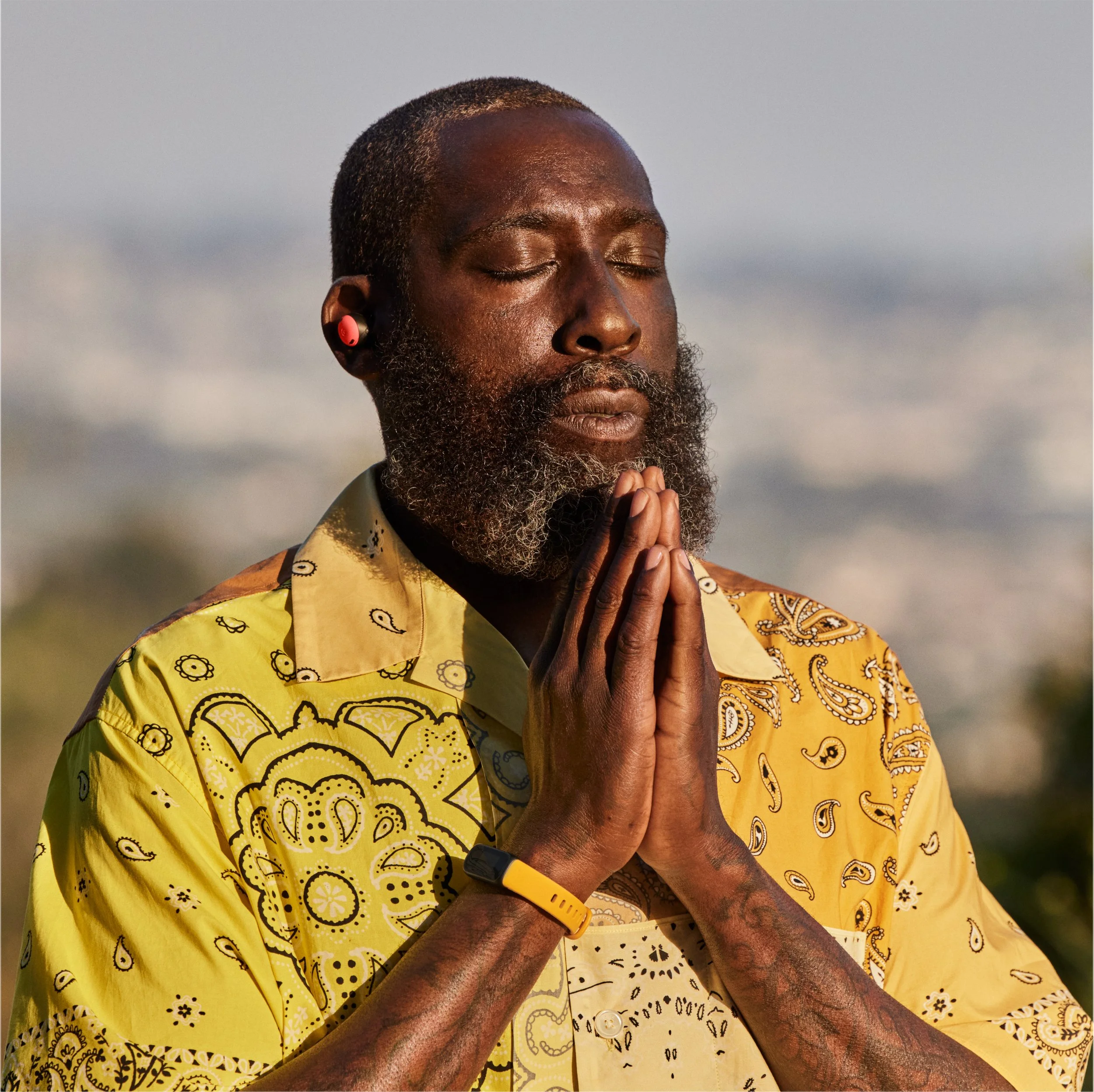 A man with a beard and closed eyes, wearing a yellow bandana and shirt with paisley patterns, stands outdoors with hands pressed together in a prayer or meditation pose, and wears a yellow wristband.