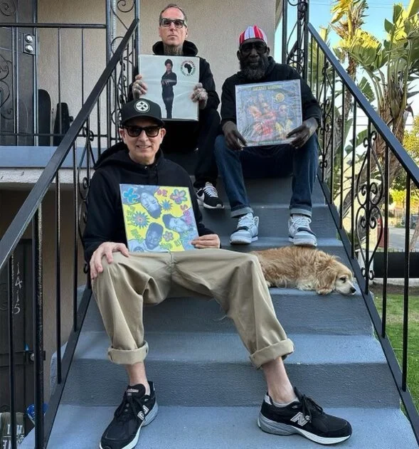 Three men sitting on stairs outside, each holding artwork, with a dog lying next to the man sitting at the bottom, all wearing casual clothing and sunglasses, with greenery in the background.