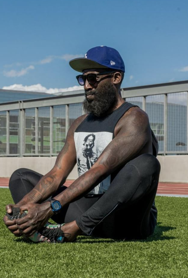 A man with a beard wearing a blue cap, sunglasses, and a black sleeveless shirt with a graphic print, sitting cross-legged on a grassy sports field holding a gaming controller.