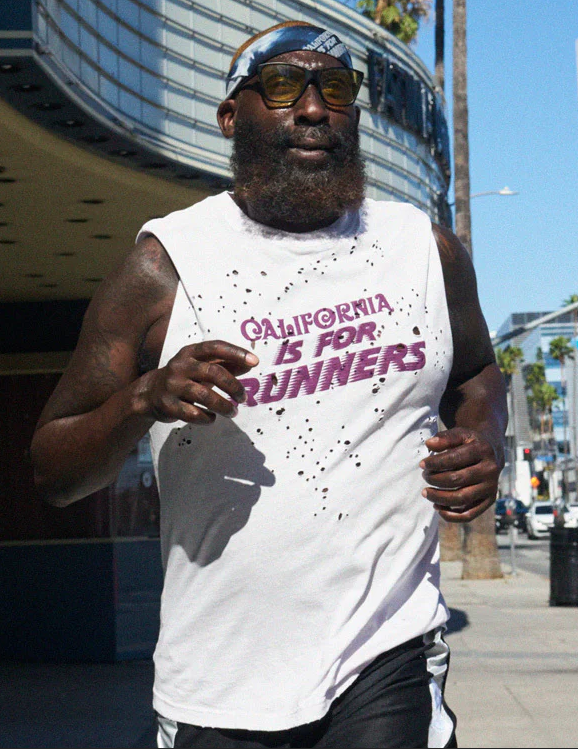 A man running outdoors, wearing sunglasses, a blue headband, a white sleeveless shirt with purple text that says 'California is for Runners,' dark shorts, and a black beard, with a city street scene in the background.