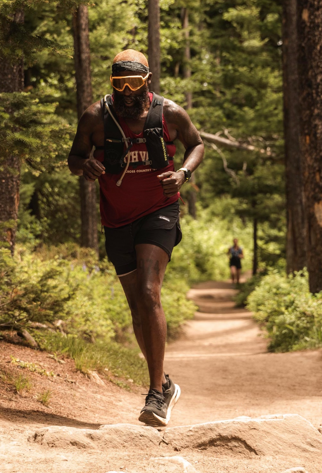 A man running on a forest trail during daytime, wearing a red tank top, black shorts, sunglasses, and carrying a hydration pack.