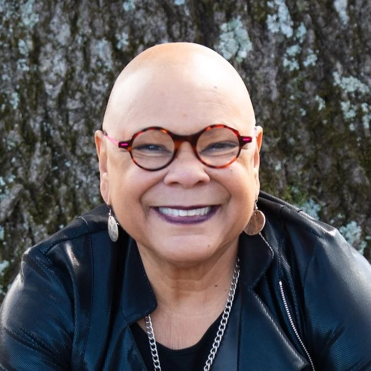 A smiling person with a shaved head, wearing round glasses, earrings, a leather jacket, and a chain necklace, standing outdoors in front of a textured tree trunk.