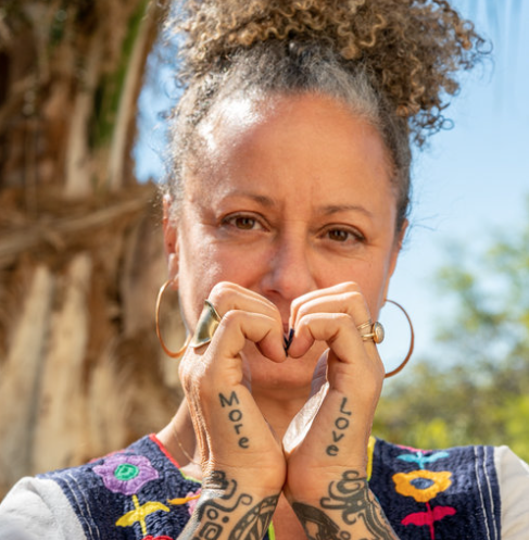 A woman with curly hair and hoop earrings making a heart shape with her hands in front of her face outdoors.