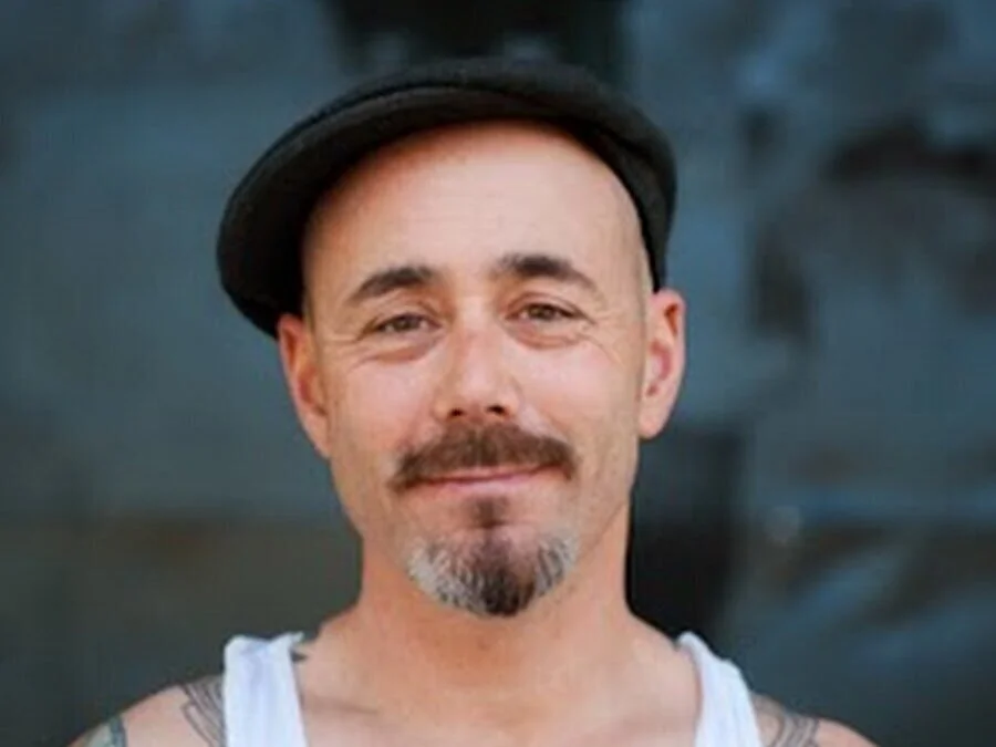 A man with a goatee and mustache wearing a black hat and a white tank top, smiling against a blurred dark background.