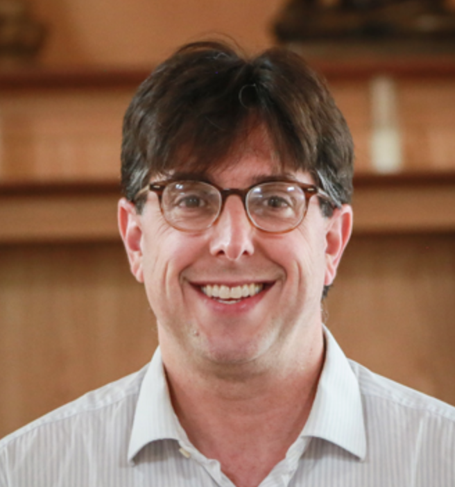 Smiling man with brown hair and glasses in a light dress shirt indoors.