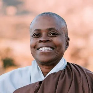 Portrait of a smiling woman wearing white and brown clothing, outdoors with blurred background.