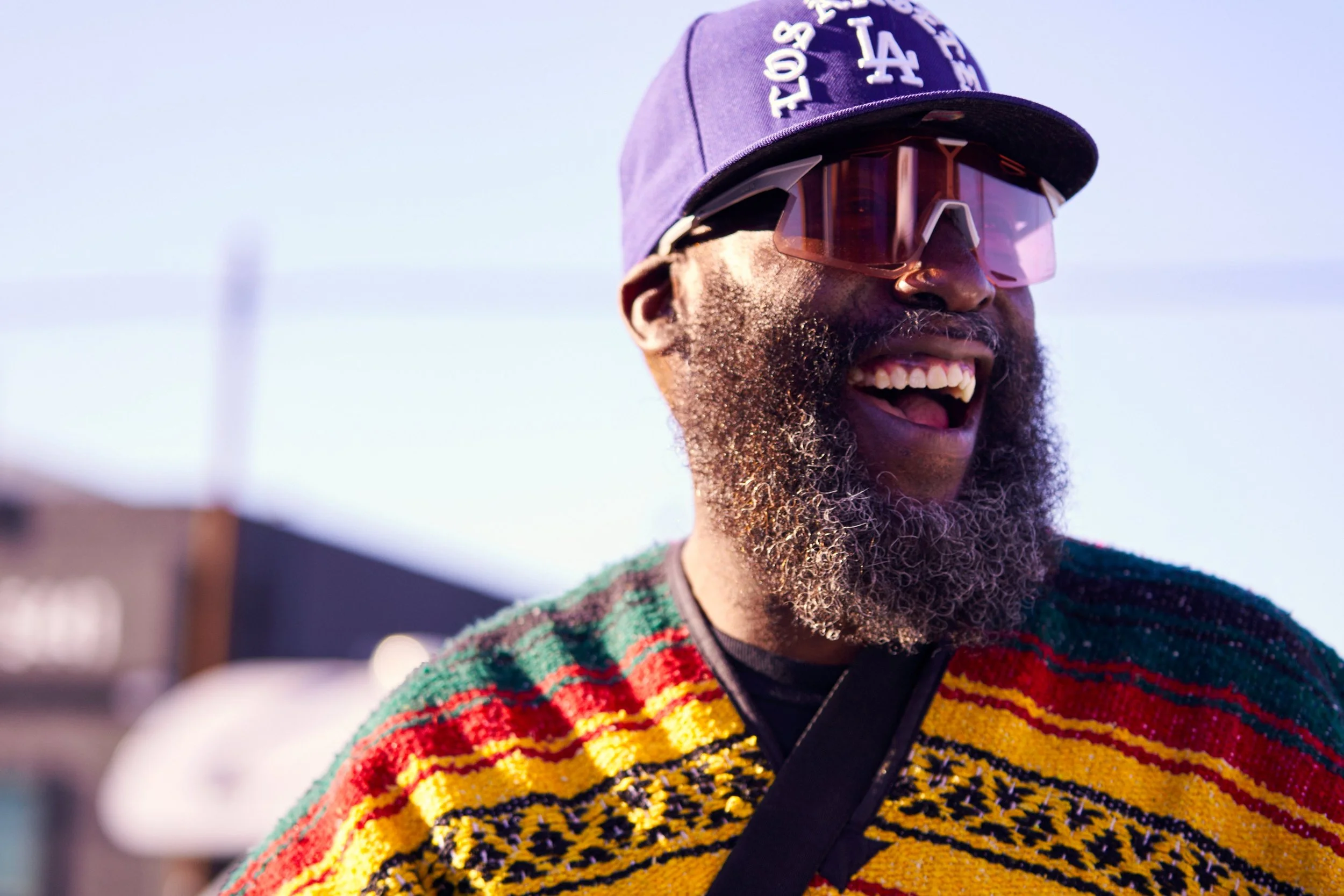 A joyful man wearing a colorful striped sweater, sunglasses, and a purple Los Angeles Dodgers hat, smiling with his mouth open, outdoors on a sunny day.