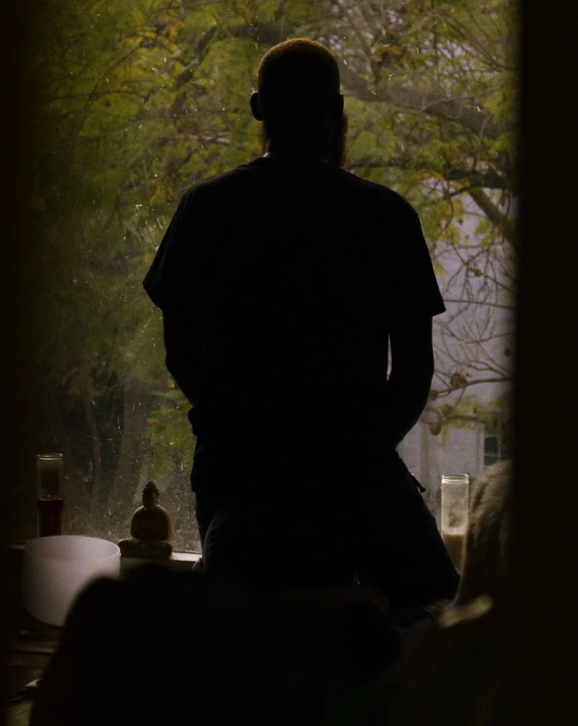 Silhouette of a person sitting by a window, overlooking a tree with autumn leaves, with candles and decorative objects on the windowsill.