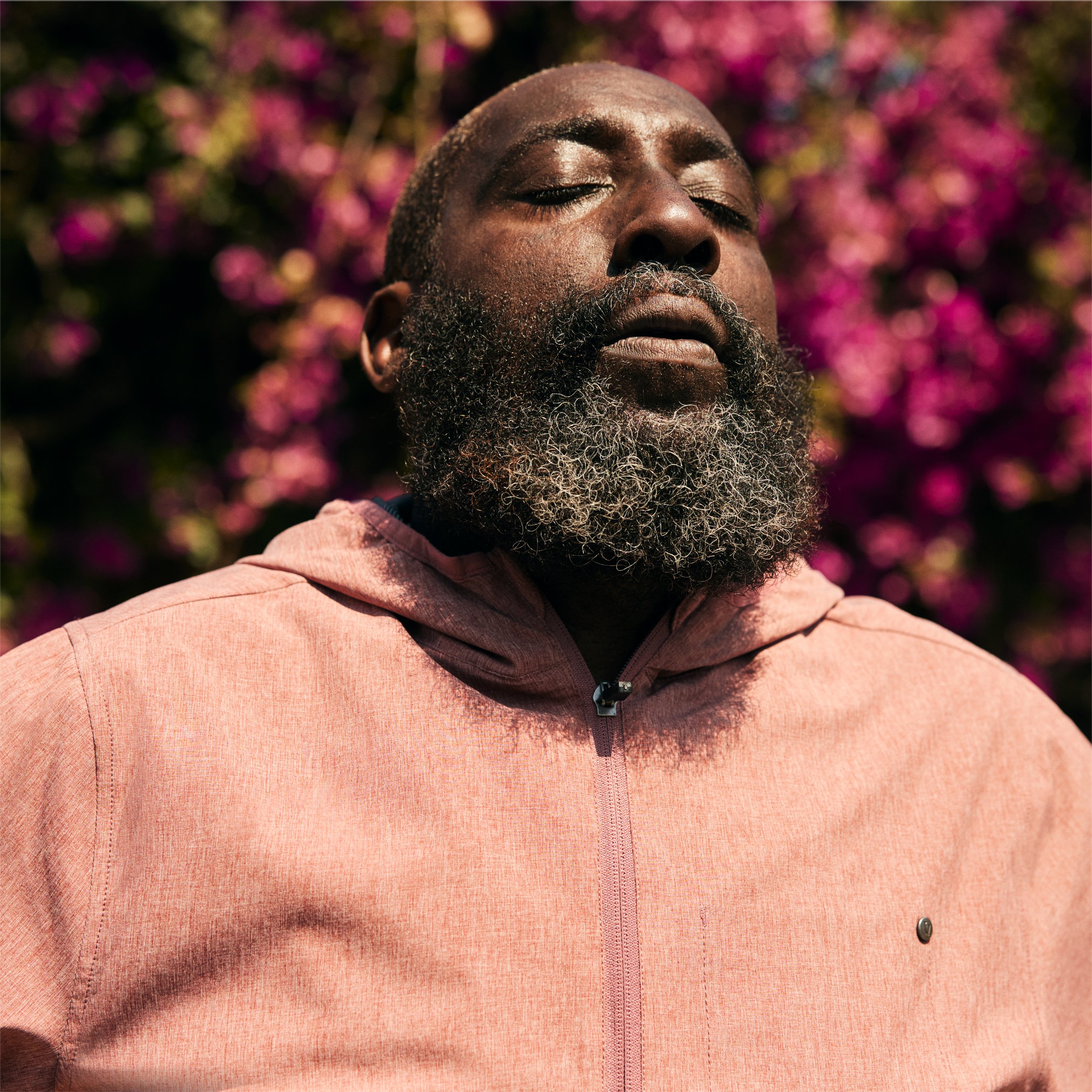 A man with closed eyes, a gray beard, and mustache, wearing a salmon-colored zip-up jacket, standing in front of pink and purple flowering bushes.