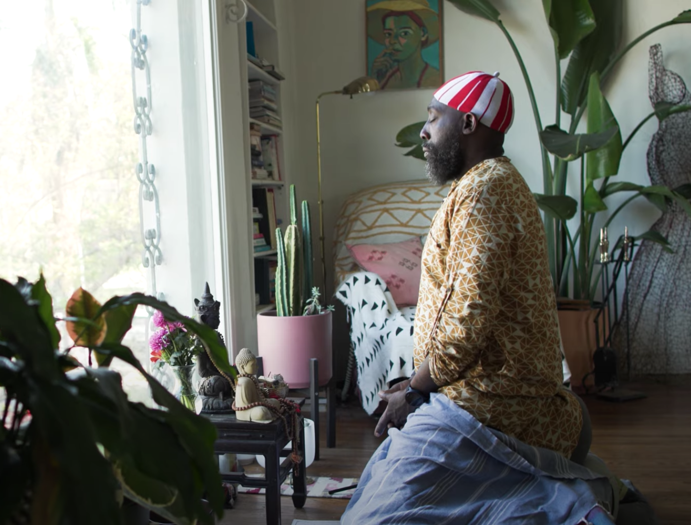 A man with dark skin, a beard, wearing a red and white striped head covering and beige patterned shirt, kneeling indoors near a prayer altar with various religious statues and flowers, practicing meditation in front of a window with natural light.