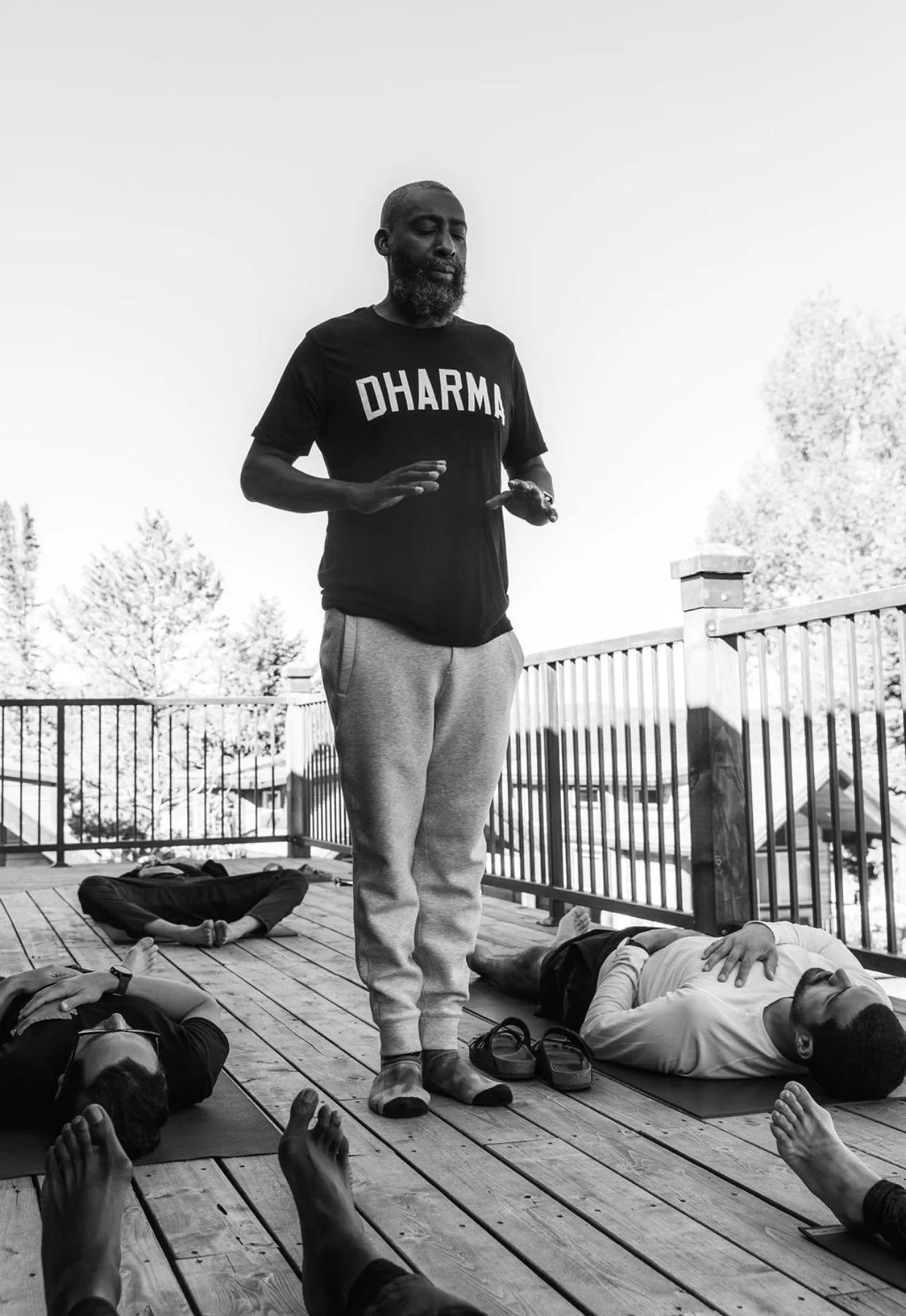 A man standing with his eyes closed in a meditative pose on a wooden deck, surrounded by people lying down with their hands raised in a yoga or meditation class outdoors, with a railing and trees in the background.