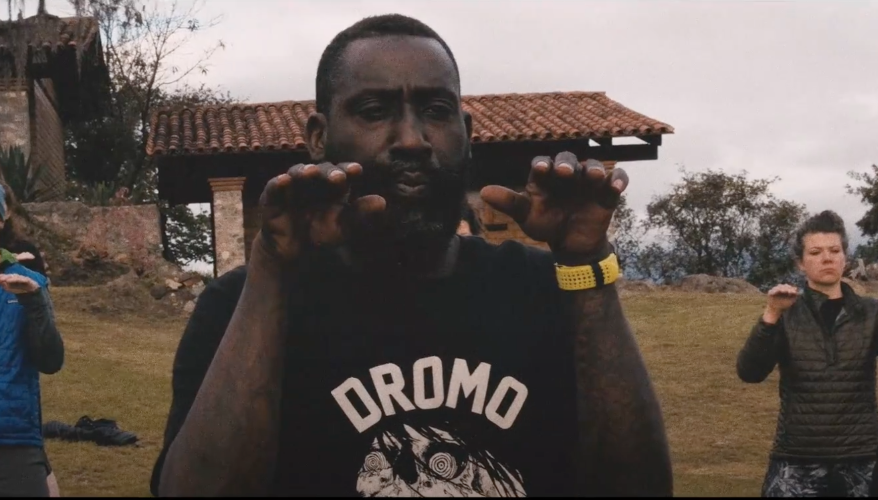 A group of people, including a man with a beard wearing a black Dromo t-shirt, outdoors during daytime with cloudy skies. The man is in the center, hands raised near his face, with other individuals around him, some in motion.