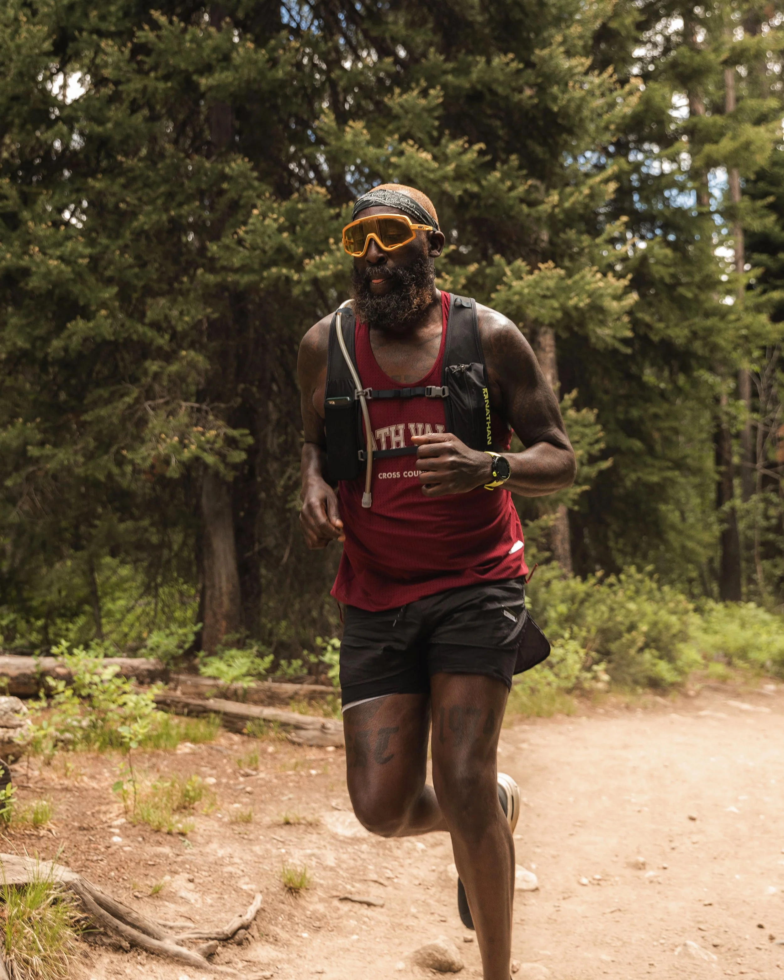 An athletic Black man with a beard running outdoors on a dirt trail surrounded by trees, wearing a red tank top, black shorts, yellow sunglasses, and carrying a hydration pack.