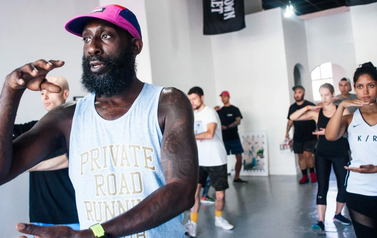 A man with a beard, wearing a pink and purple cap, a light blue tank top, and a yellow watch, appears to be leading a fitness or dance class with a group of diverse people in a spacious room with white walls and large windows.