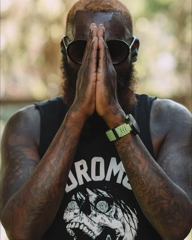 Man with tattoos, wearing sunglasses, a black sleeveless shirt with a skull graphic and the text 'PROM,' a green wristwatch, with hands pressed together in a prayer or meditative pose.