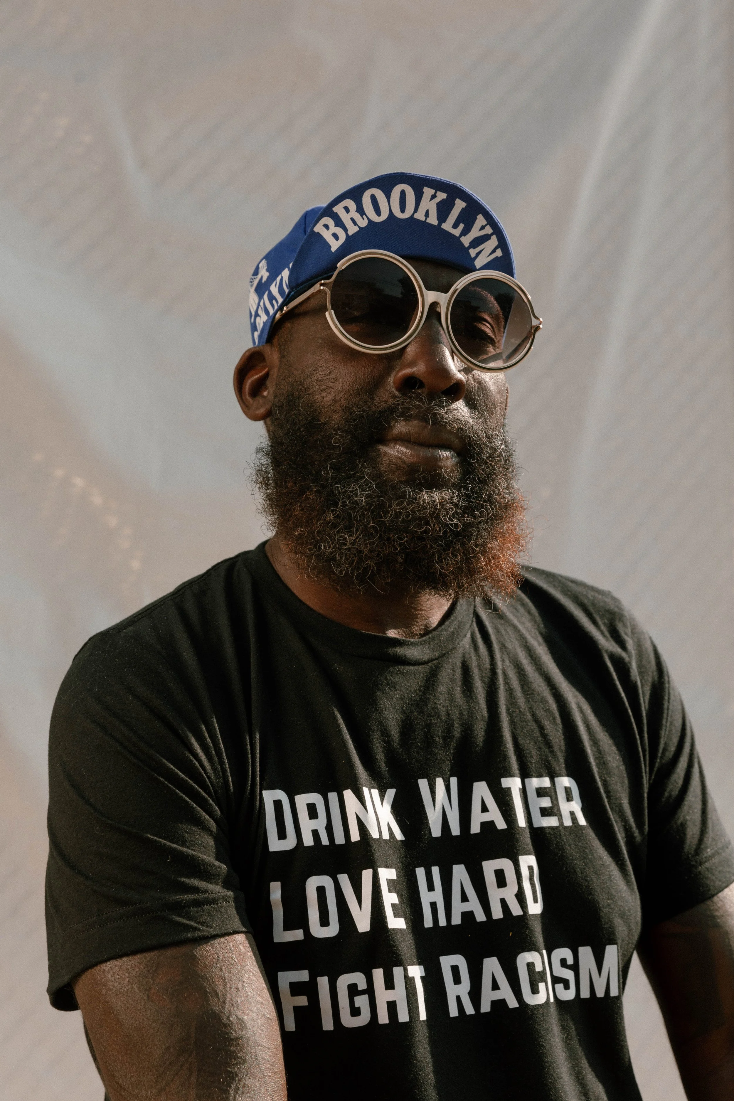 Close-up of a man with dark skin, beard, and sunglasses wearing a blue headband that says 'Brooklyn' and a black T-shirt with white text promoting anti-racism and love.