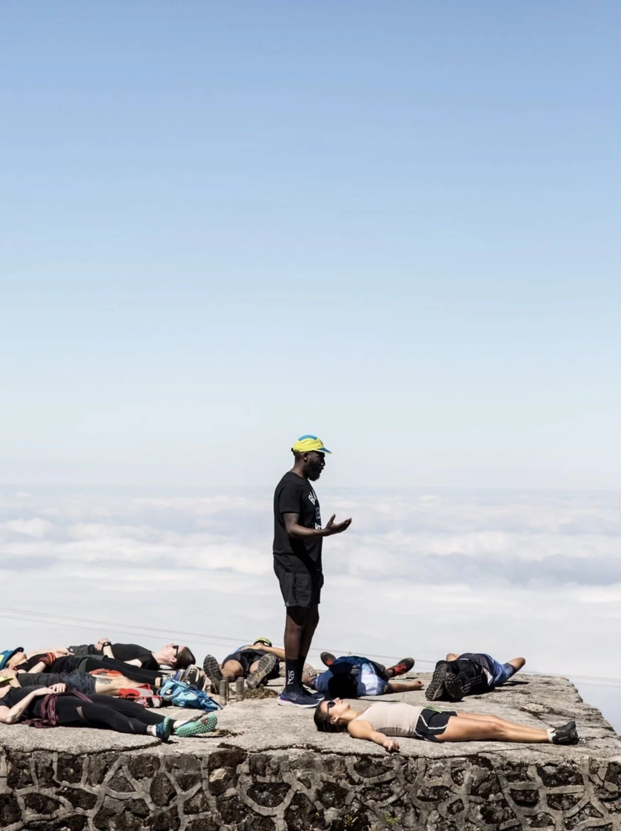 A man standing on a stone ledge with his hands up, surrounded by people lying resting on the ground. The background features a sky filled with clouds and above the clouds, indicating they are at a high altitude.