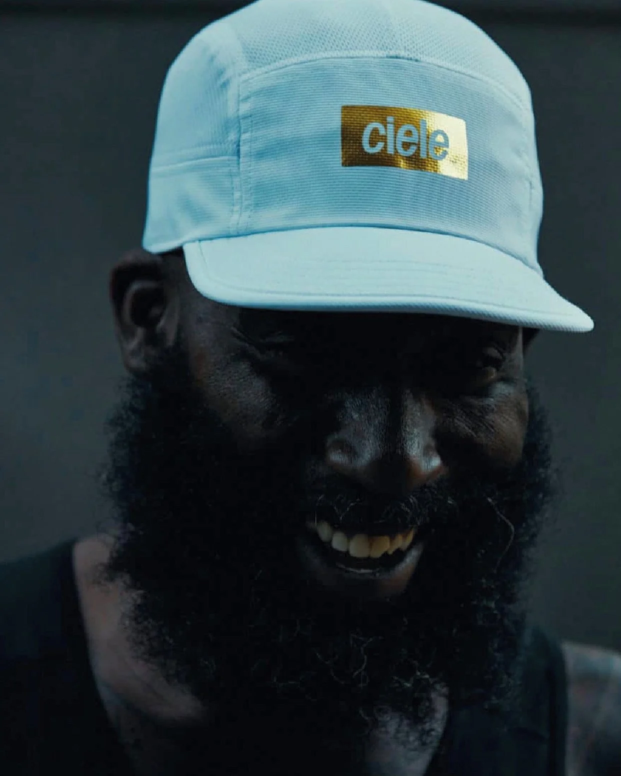 Person with dark skin, black beard, and mustache smiling, wearing a white cap with a gold and blue "ciele" logo.