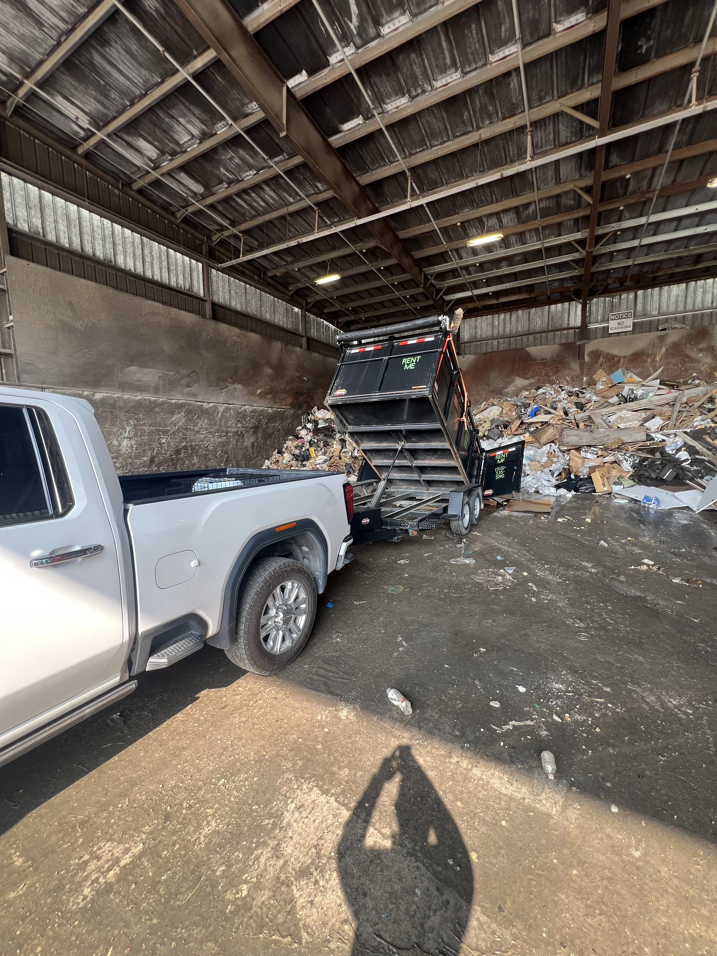 A large black dumpster is tipped over and overflowing with trash, spilling debris and waste onto the ground. A silver pickup truck is parked nearby, and the area appears to be a trash or waste disposal site with a muddy, dirty floor and a high metal roof overhead.