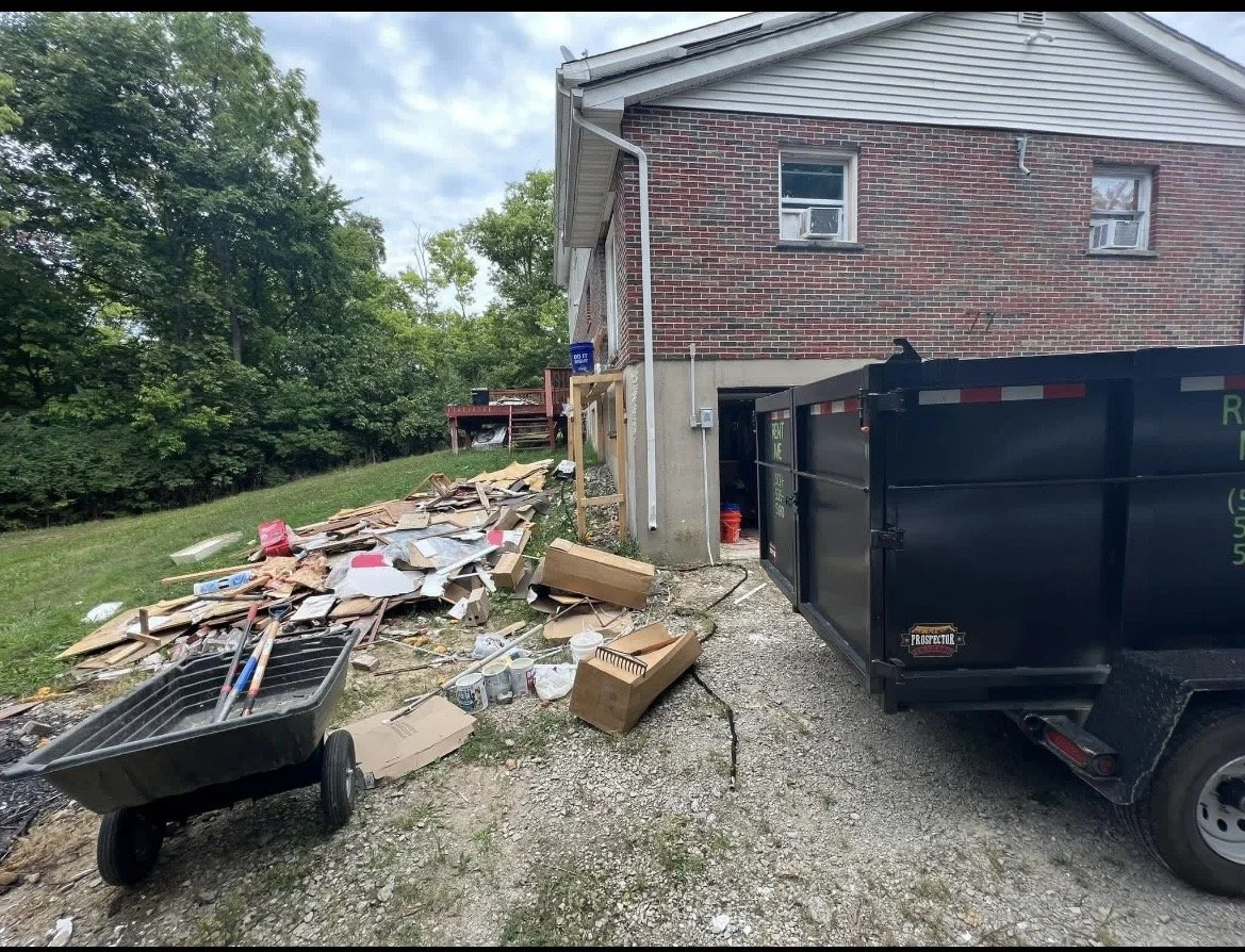 Side of a house with construction and yard waste, including cardboard, debris, a wheelbarrow, and a large black dumpster.