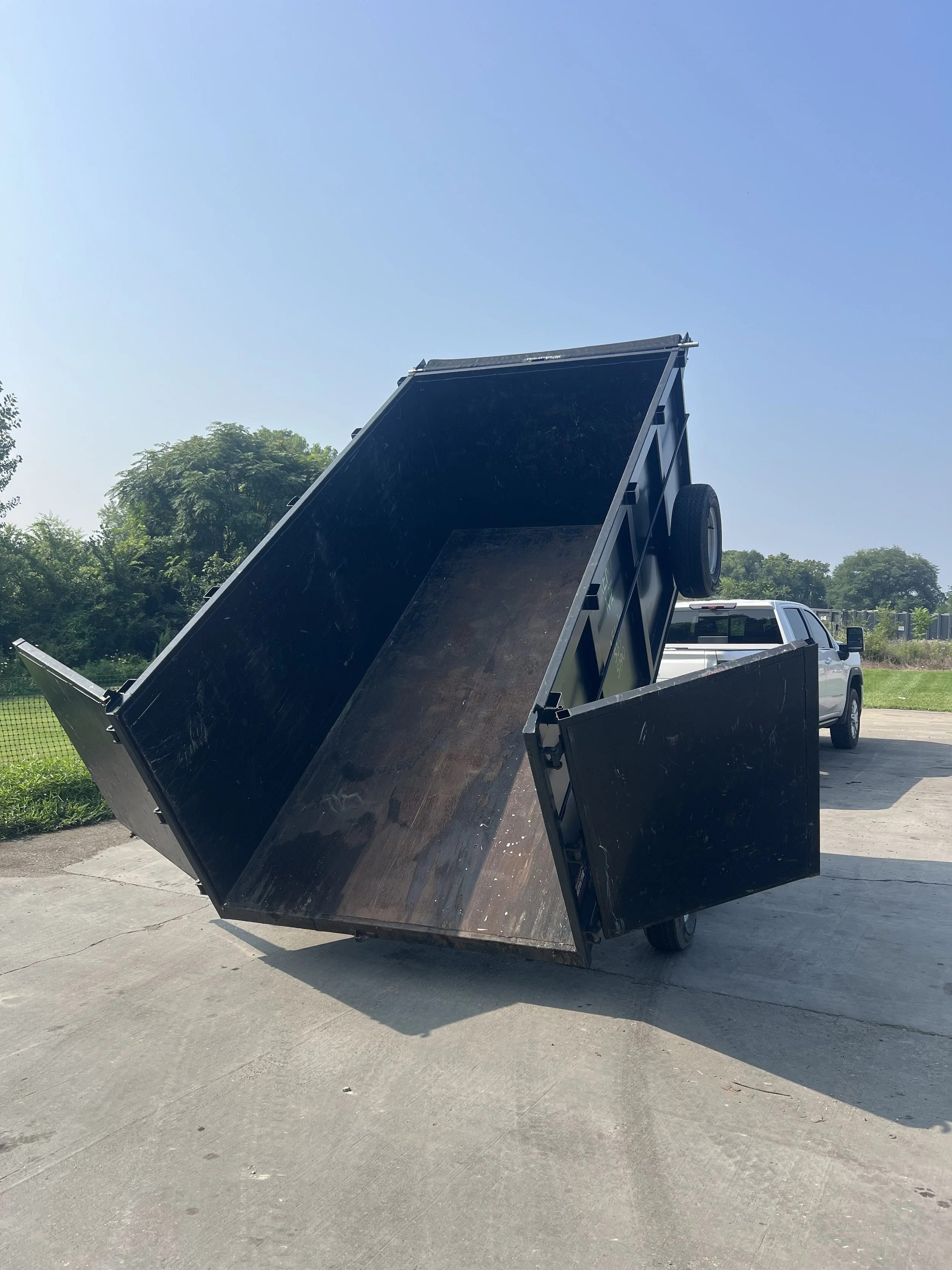 A dump truck with its bed raised, in a parking lot next to trees and a grassy area under a clear blue sky.