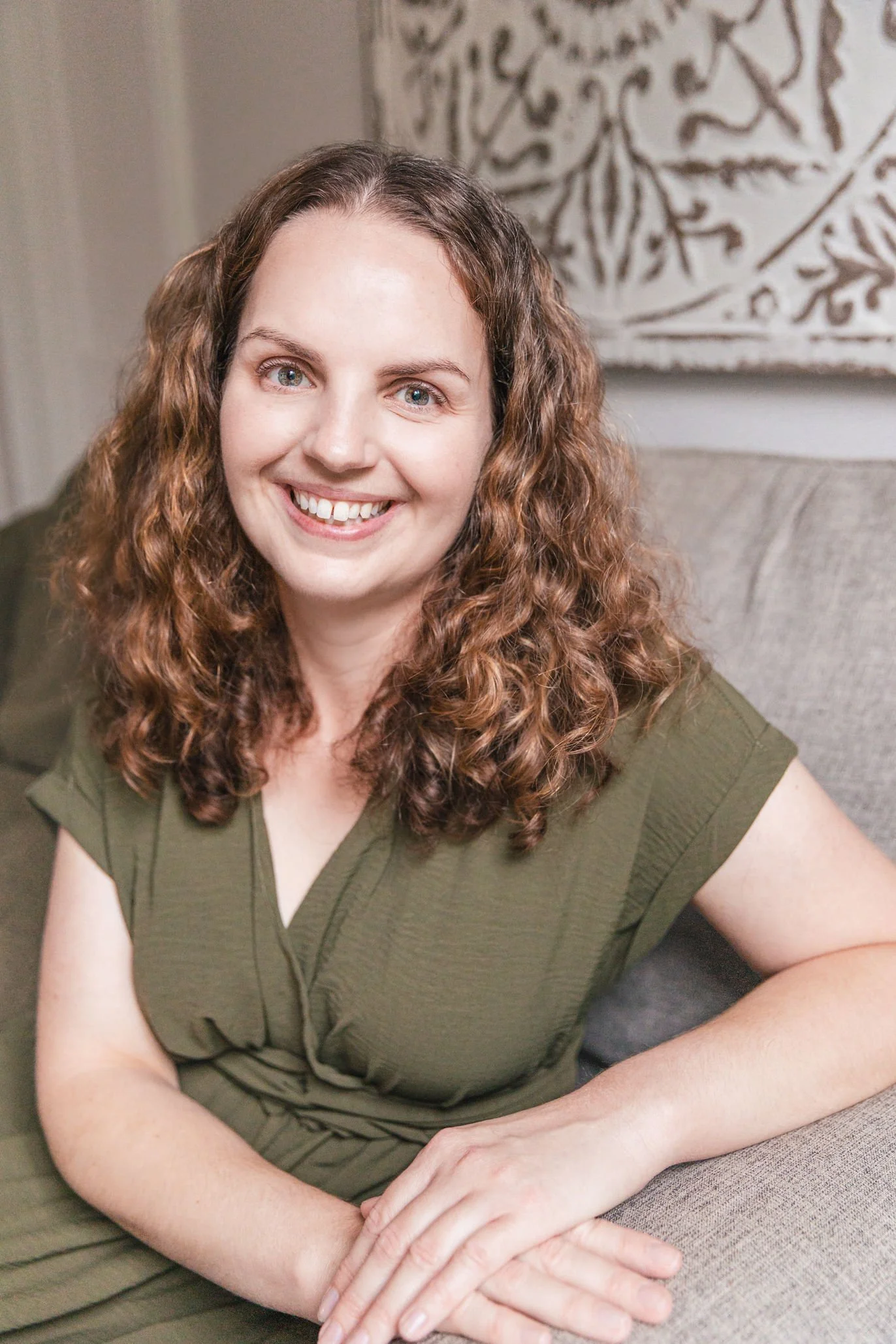 A woman with long curly brown hair and blue eyes sitting on a gray couch, wearing an olive green top, smiling at the camera with her hands resting on her lap. Behind her is a decorative wall hanging with a floral pattern.