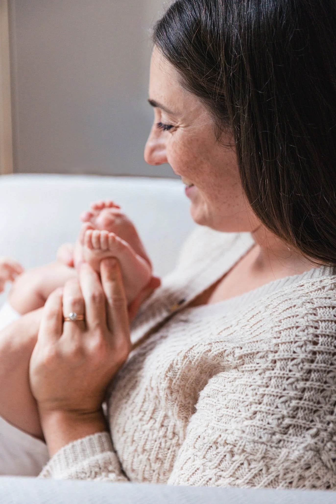 Close-up of a woman with dark hair and freckles holding a baby's hand, both appearing to be in a tender moment indoors.