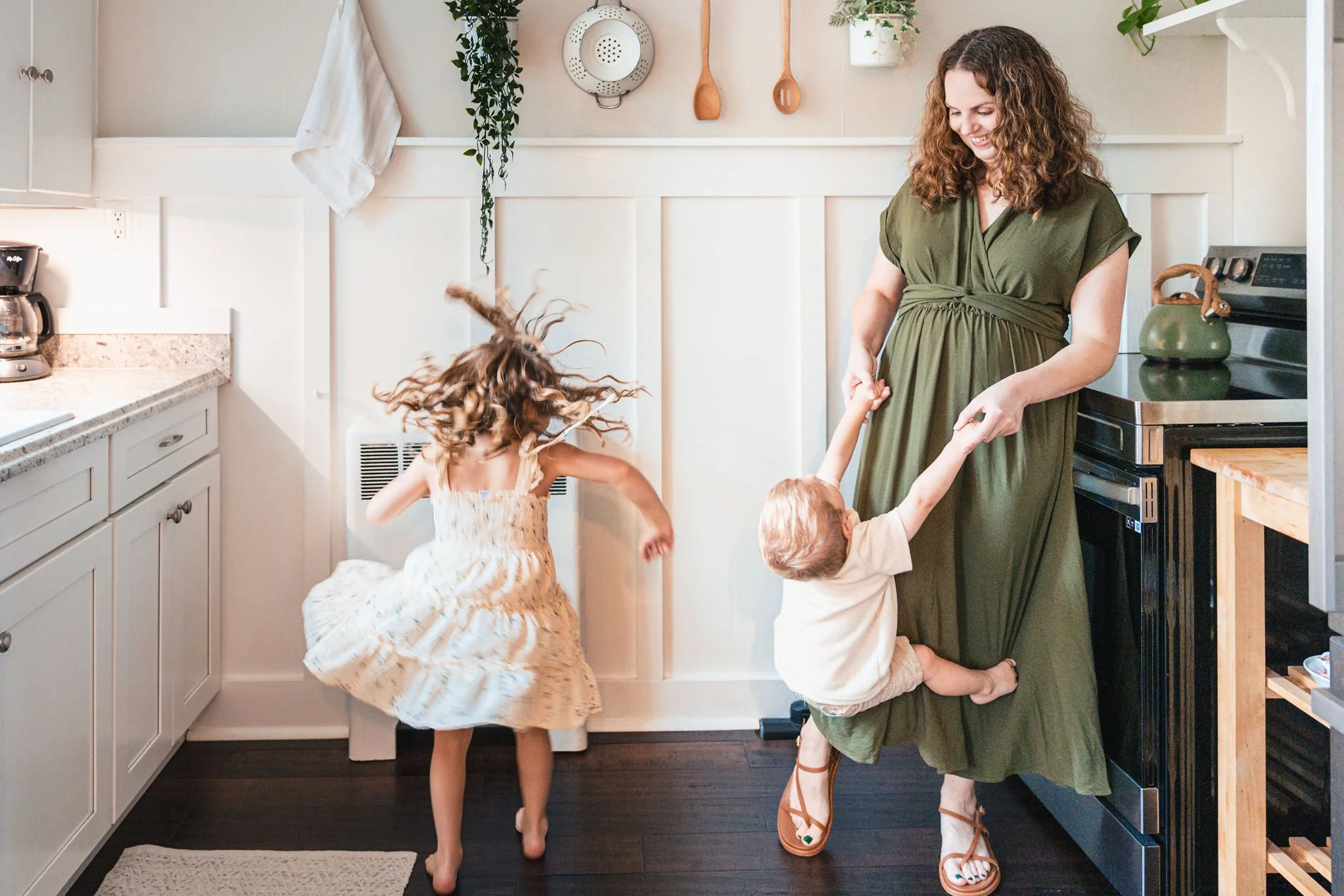 A mother in a green dress playing with two young children in a kitchen, with one child spinning and the other holding her hand.
