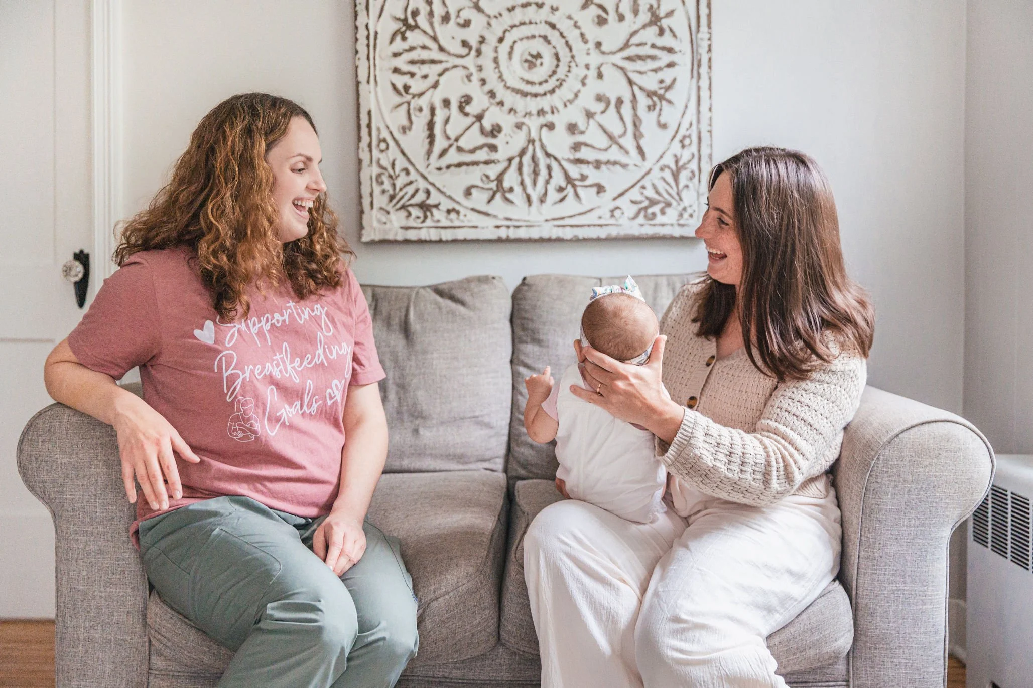 Two women with curly and straight brown hair, smiling and engaging with a baby who is sitting on one woman's lap, in a cozy living room with a decorative wall hanging.