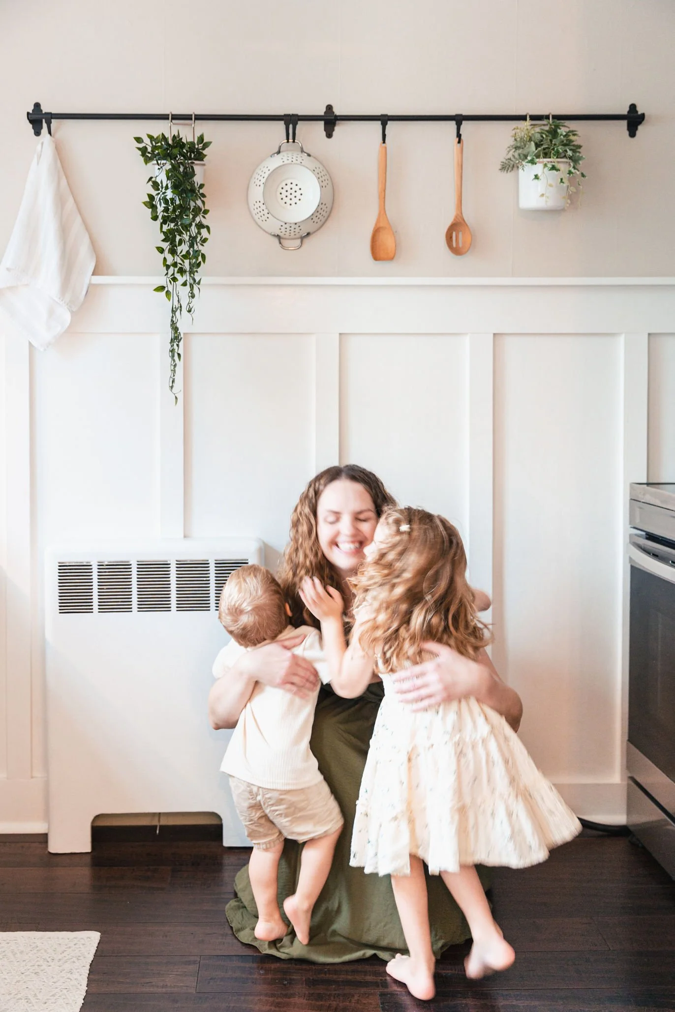 A woman with curly hair sitting on the floor, hugging and surrounded by children, smiling and embracing in a home setting with a white wall and kitchen appliances in the background.