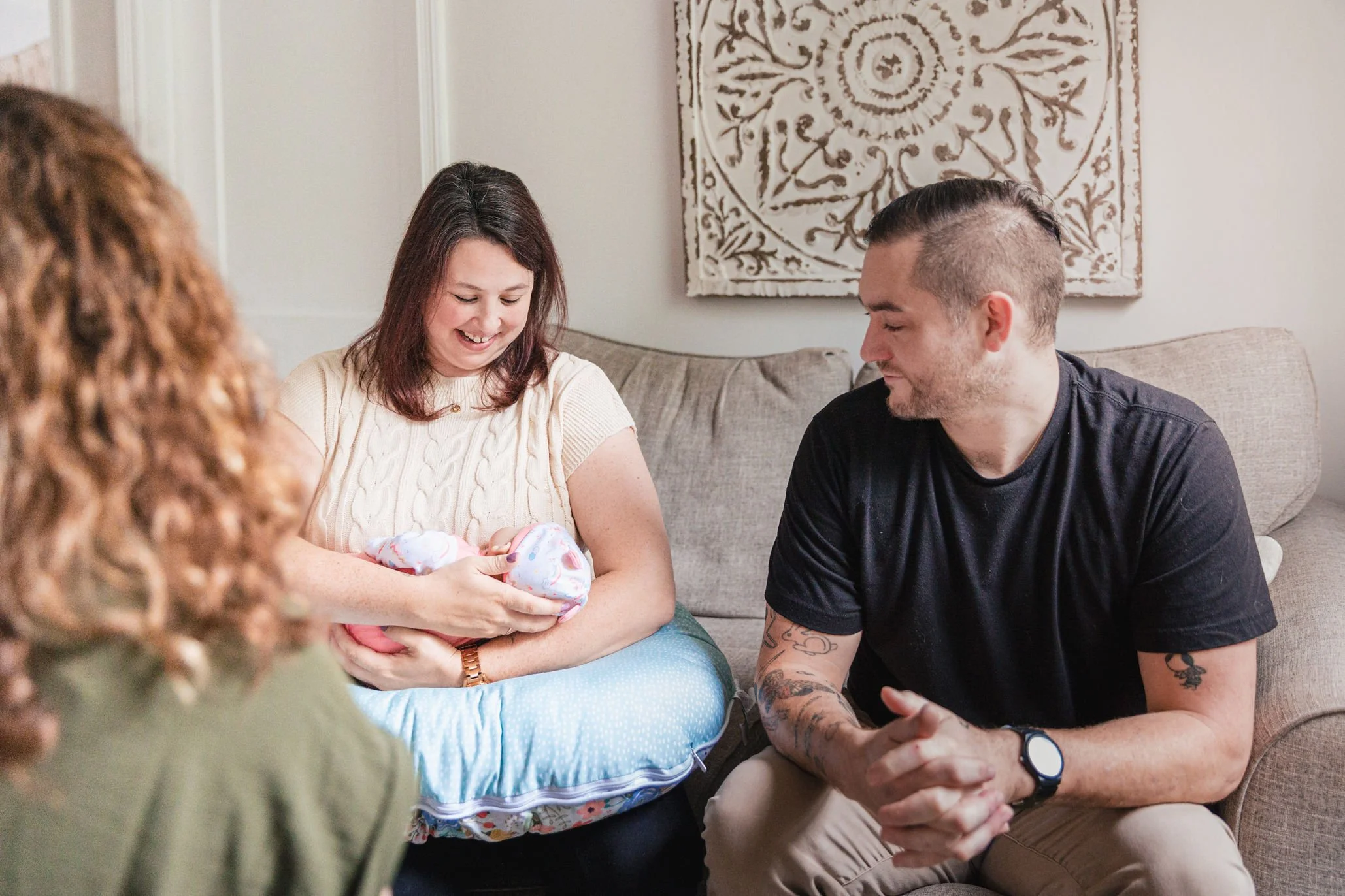 A pregnant woman holding a baby doll wrapped in a colorful blanket while a man with tattoos and glasses sits beside her, in a cozy living room with beige couch and decorative wall art learning about how to feed their baby.