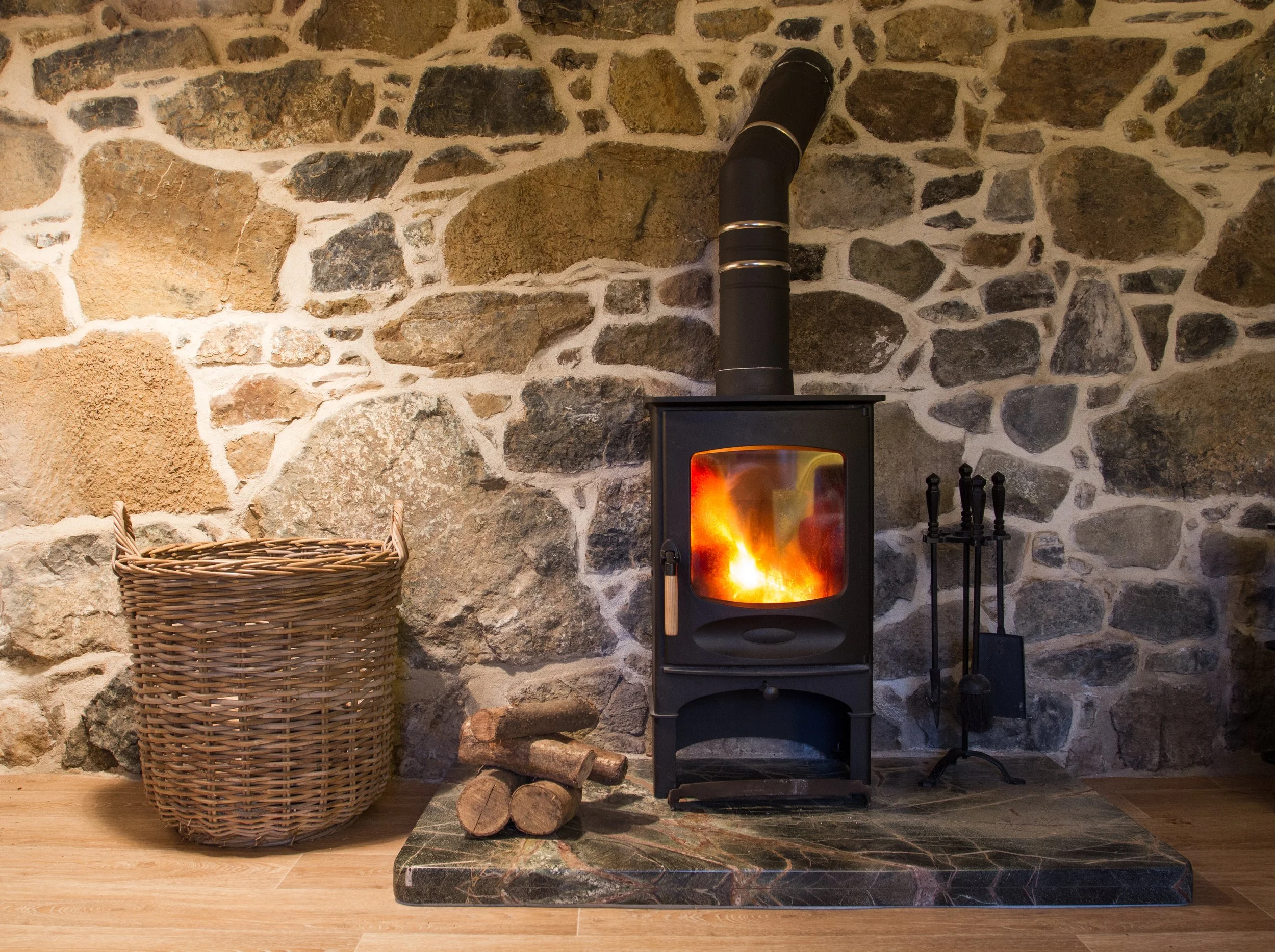 A stone fireplace with a wood stove burning inside. To the left, a large woven basket and some logs are on the floor. To the right, fireplace tools are standing next to a marble hearth.