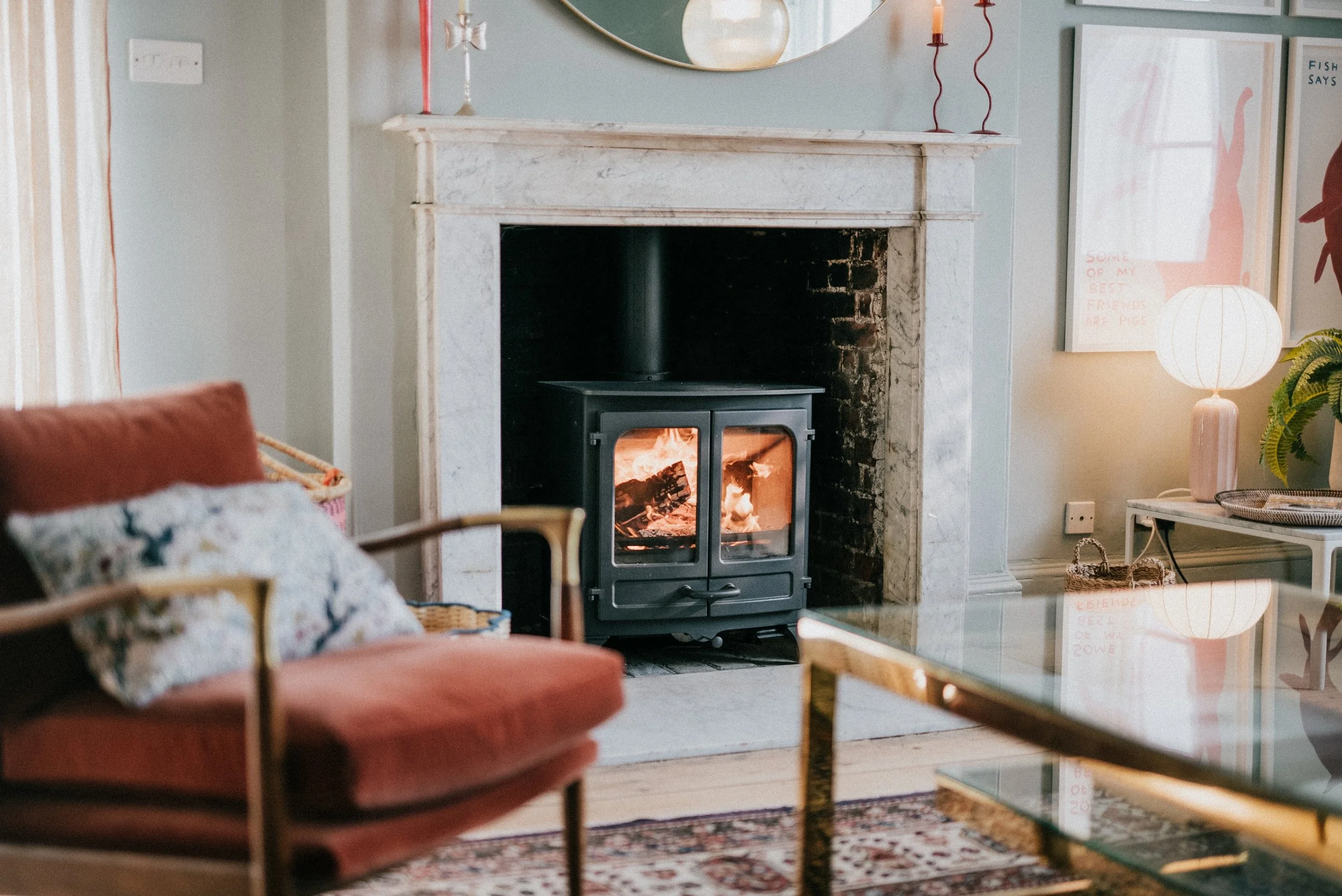 Living room with white marble fireplace, black wood stove, pink armchair with floral cushion, paper lantern lamp, and framed artwork on the wall.