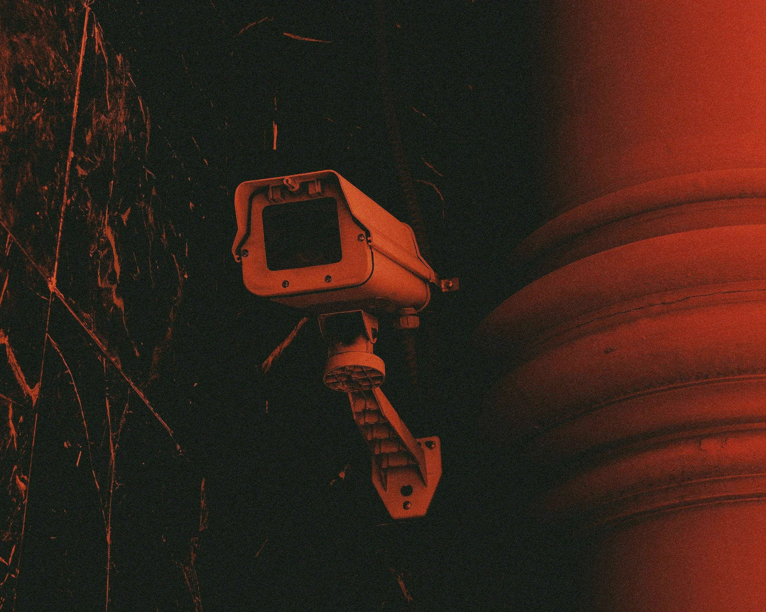 A traffic camera mounted under a concrete bridge structure, illuminated by orange-red lighting, with some branches or plant matter visible on the left side.