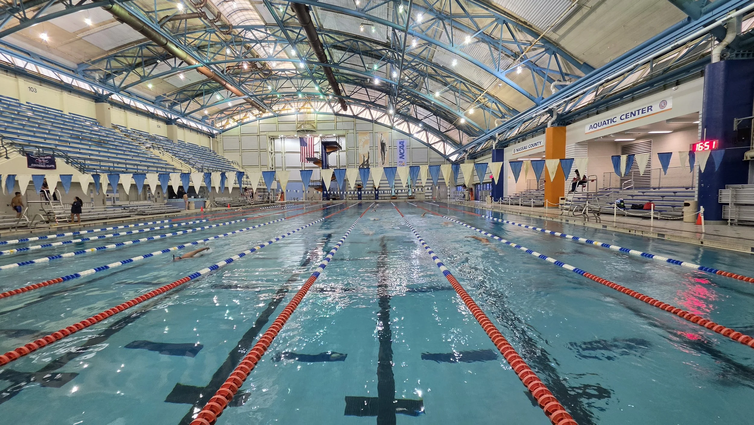 Indoor swimming pool with lanes, swimmers, and spectators. Elevated seating on the sides and an American flag in the background.