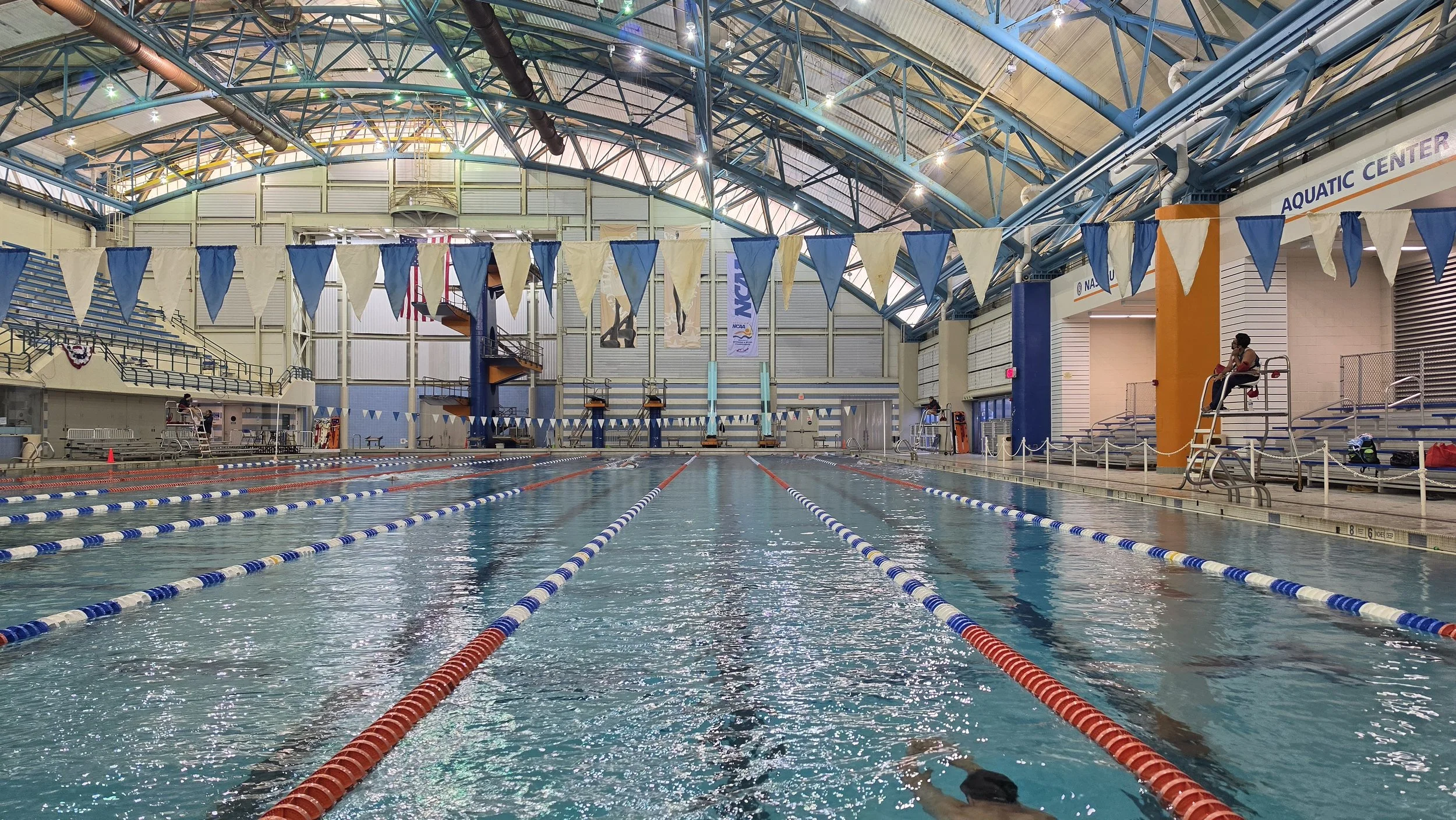 Indoor swimming pool with lane dividers, starting blocks, and swimmers, under a high arched ceiling with blue support beams.