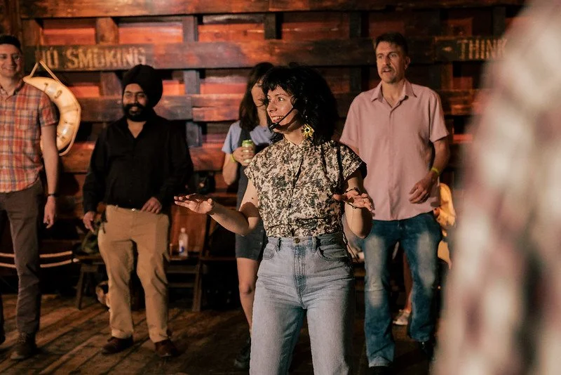A woman, wearing a vintage floral blouse and jeans, has a mic in a warmly lit room with a wooden background. Several people stand behind her, watching and listening, with some holding drinks while learning to dance.