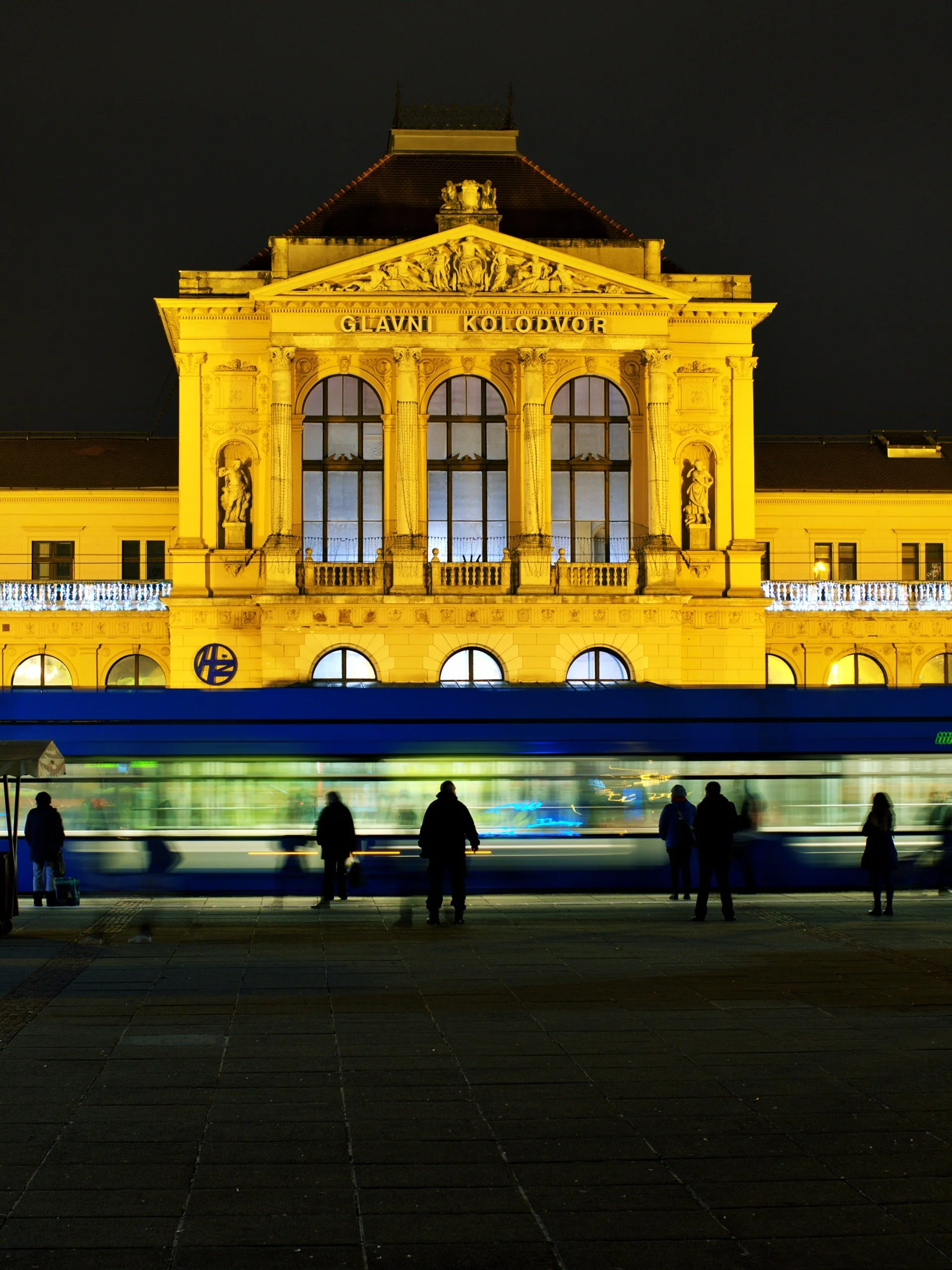 Night view of a historic building with illuminated facade, large arched windows, and statues, with several people standing on the sidewalk and a blurred passing bus in front.