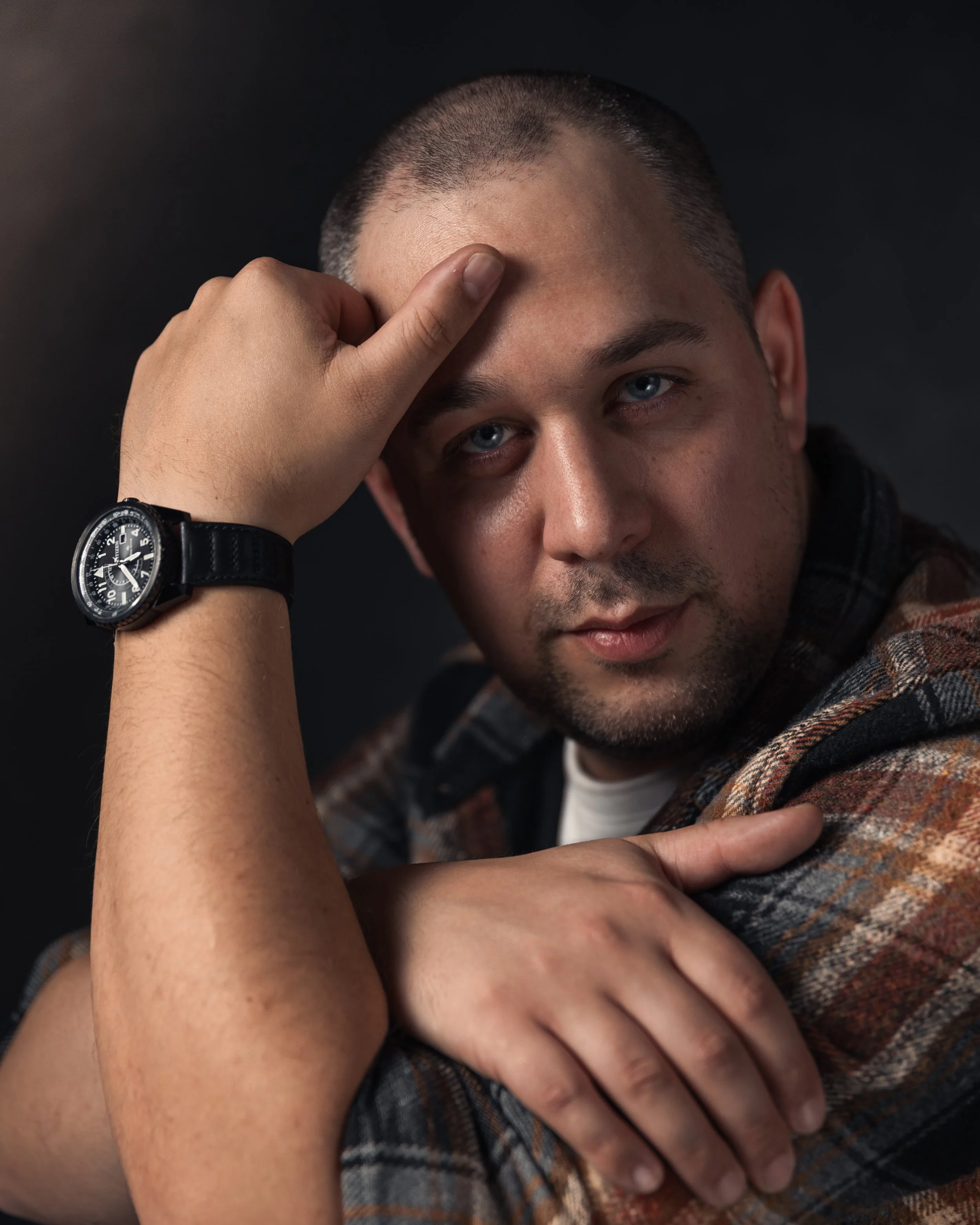A man with blue eyes and short hair, wearing a plaid shirt and a wristwatch, rests his head against his arm while touching his forehead with his finger, against a dark background.