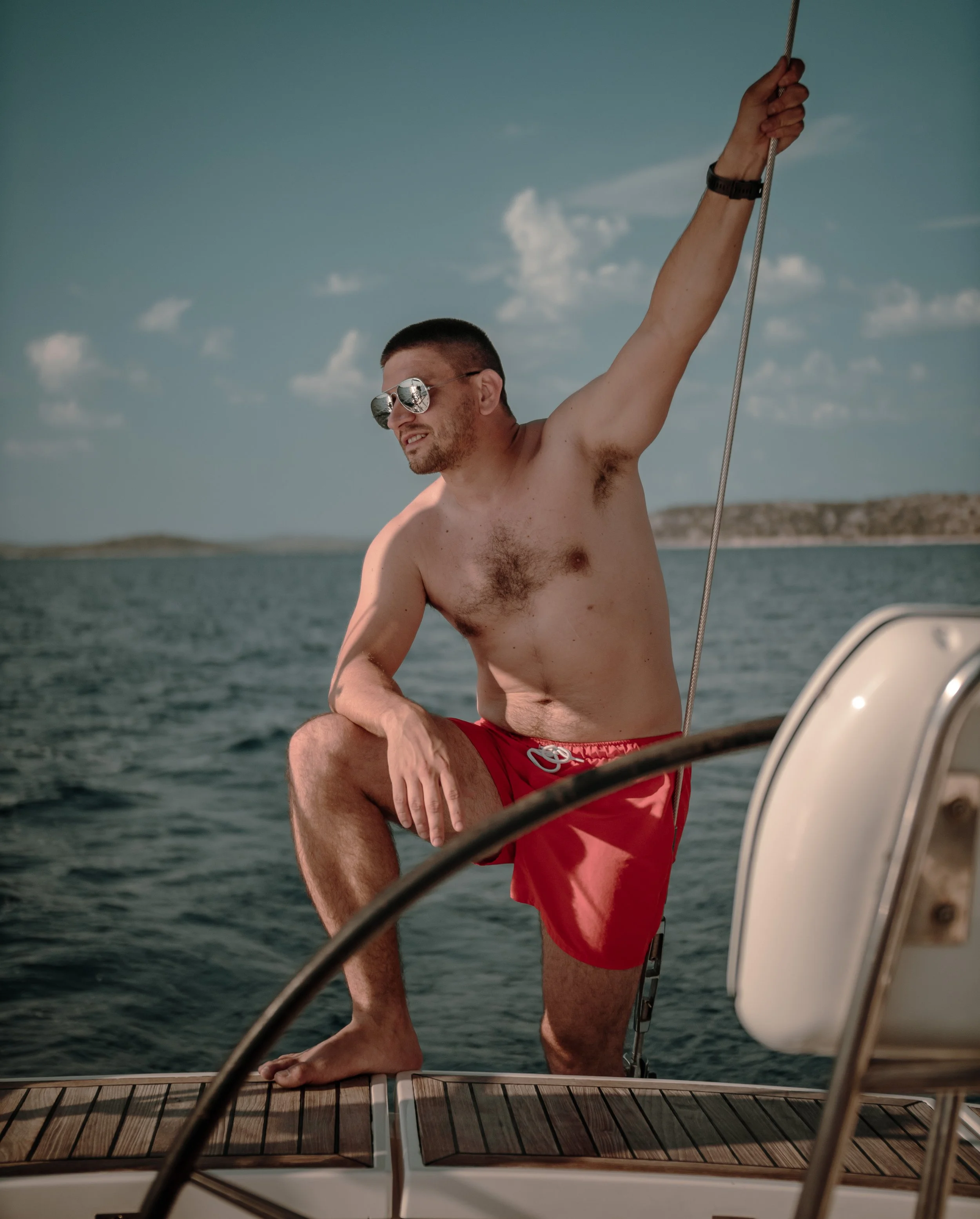 A shirtless man wearing red shorts and sunglasses sitting on a boat, holding a rope, with water and a distant shoreline in the background.