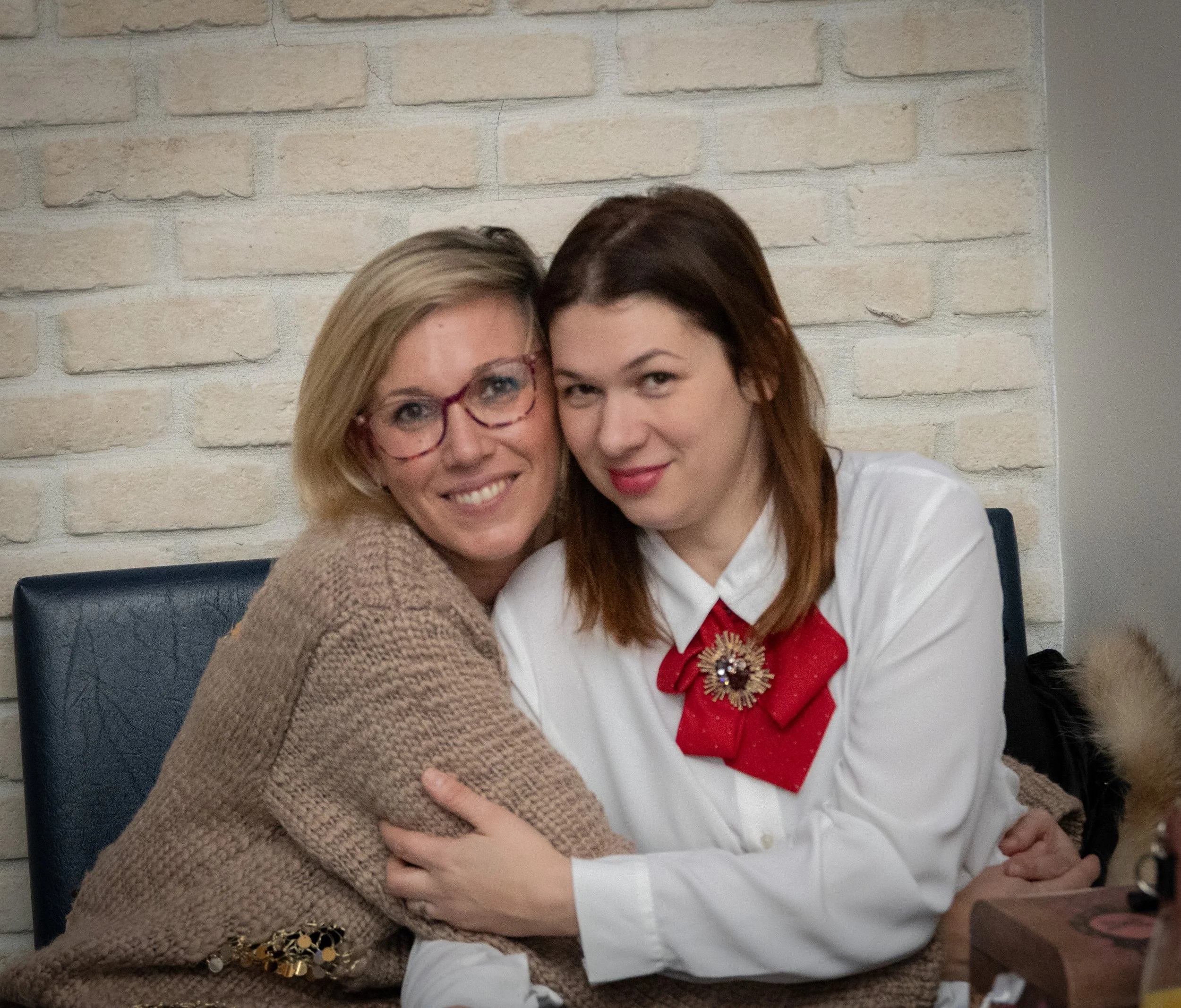 Two women, one older with blonde hair and glasses, and one younger with red hair, hugging and smiling in front of a brick wall.