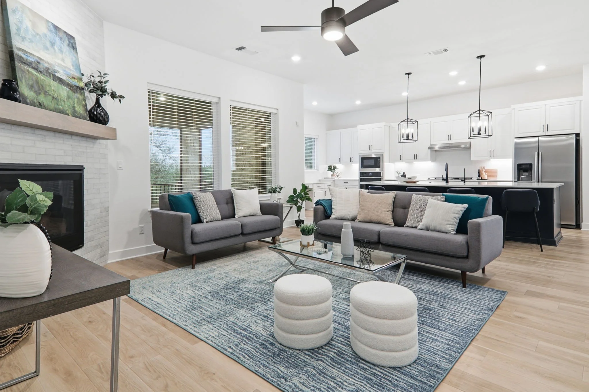 Modern open-plan living room and kitchen with gray sofas, a blue rug, white cabinets, stainless steel appliances, black pendant lights, and large windows with blinds.