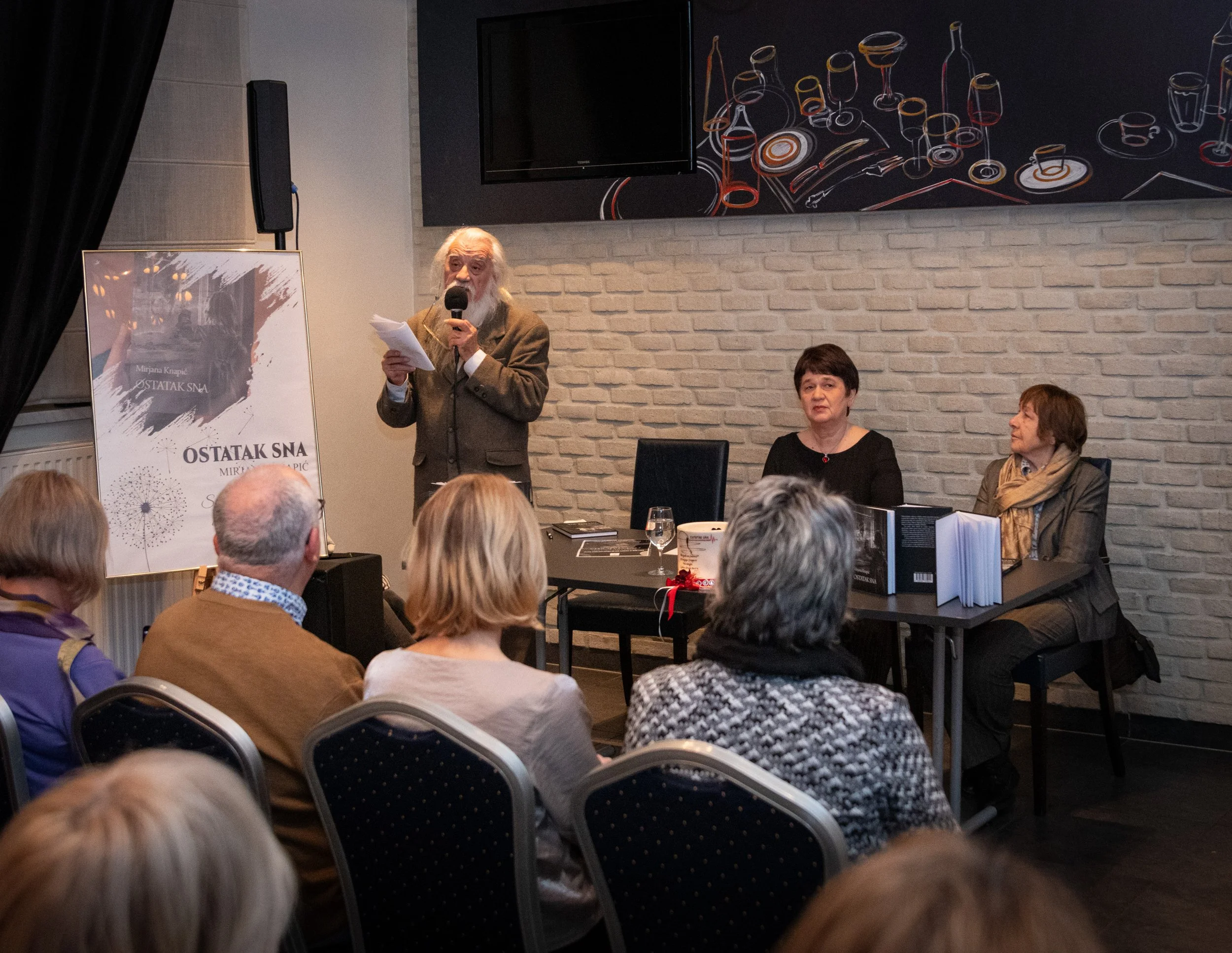 An elderly man with a white beard and hair speaks into a microphone during a presentation or event, with two women seated at a table beside him. Audience members are visible in the foreground.
