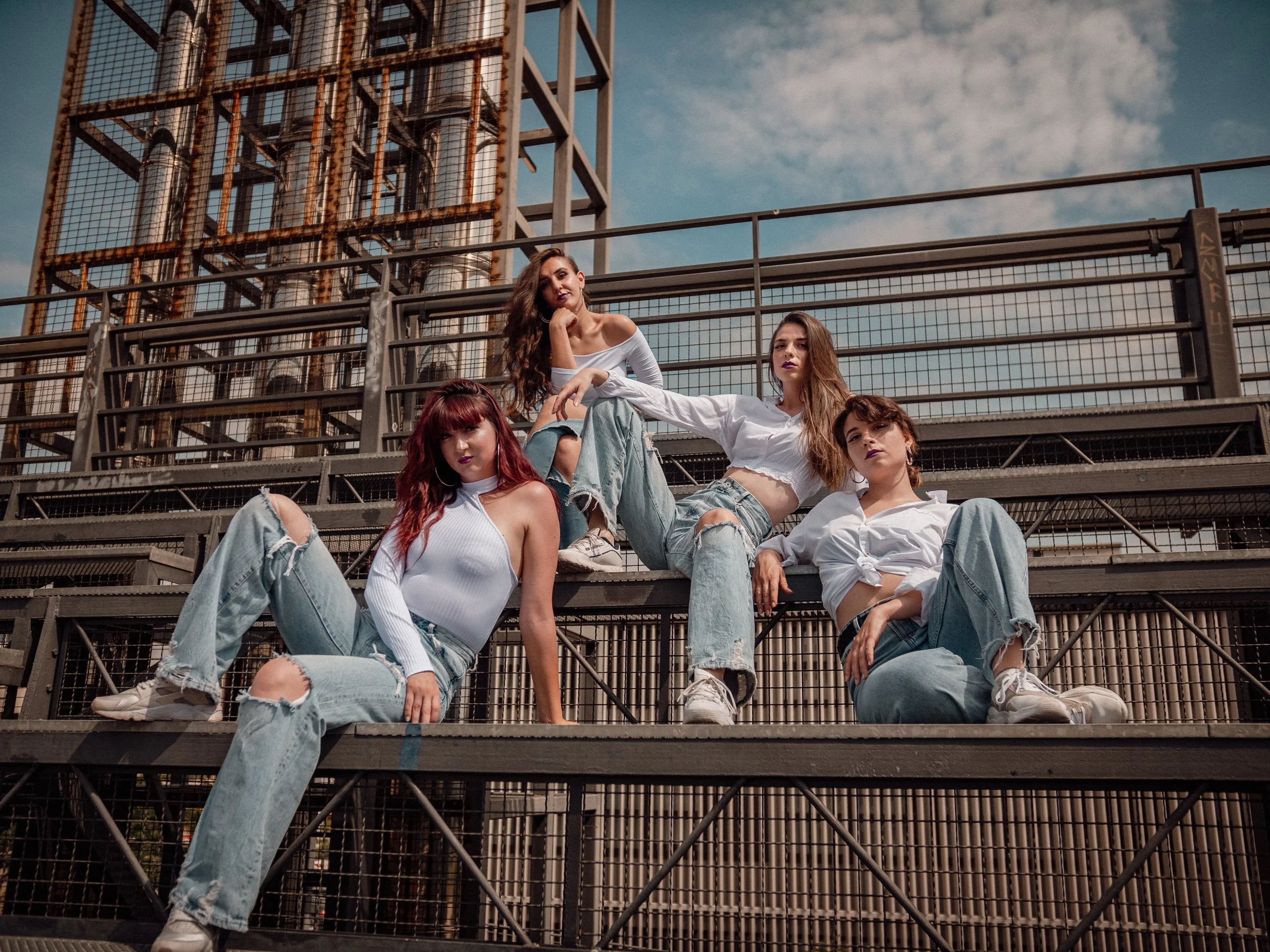 Four young women sitting on metal bleachers at an outdoor location with a large industrial structure in the background under a partly cloudy sky.