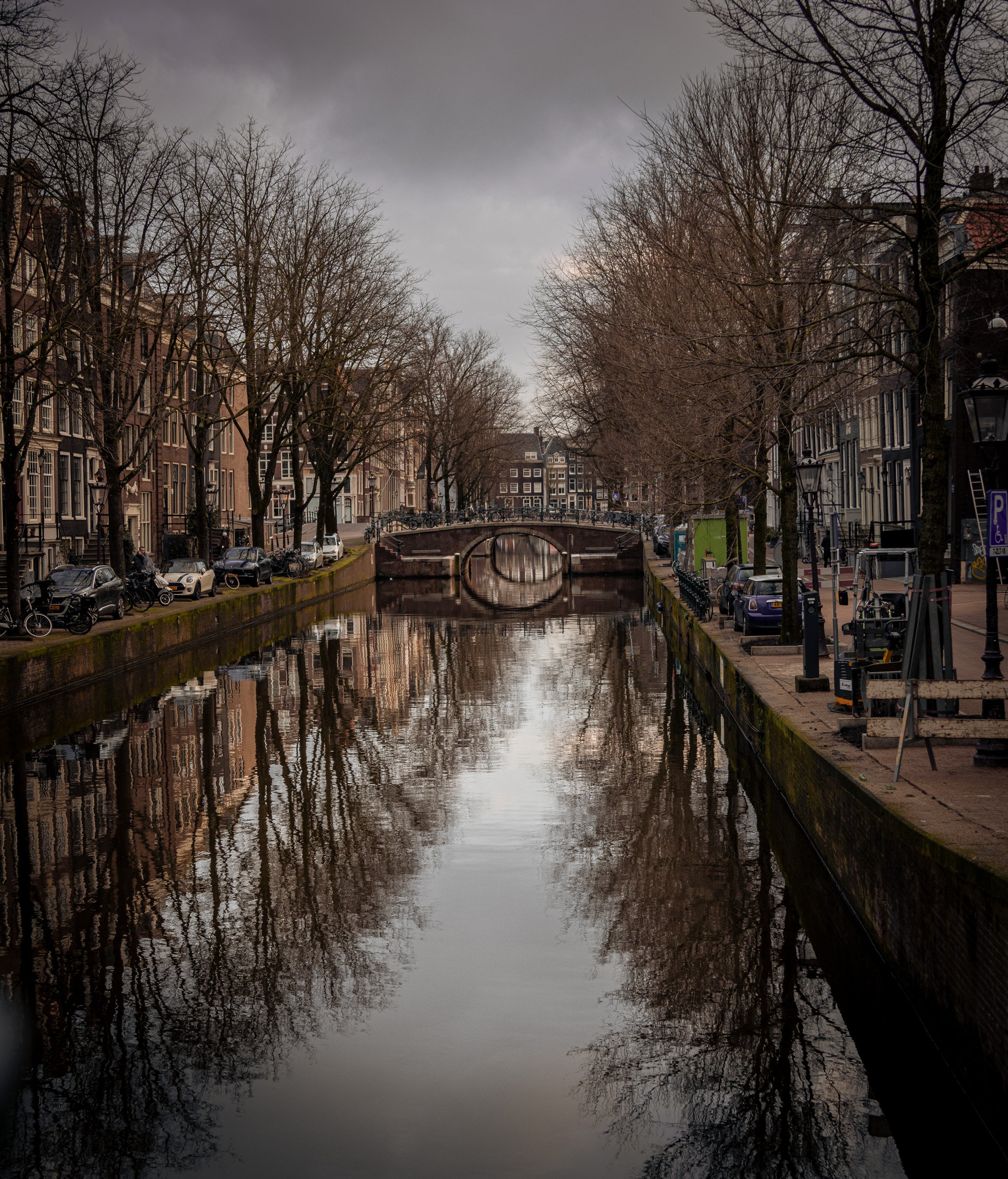 A canal lined with leafless trees and traditional European buildings under a cloudy sky, with reflections in the water and bicycles parked along the sides.