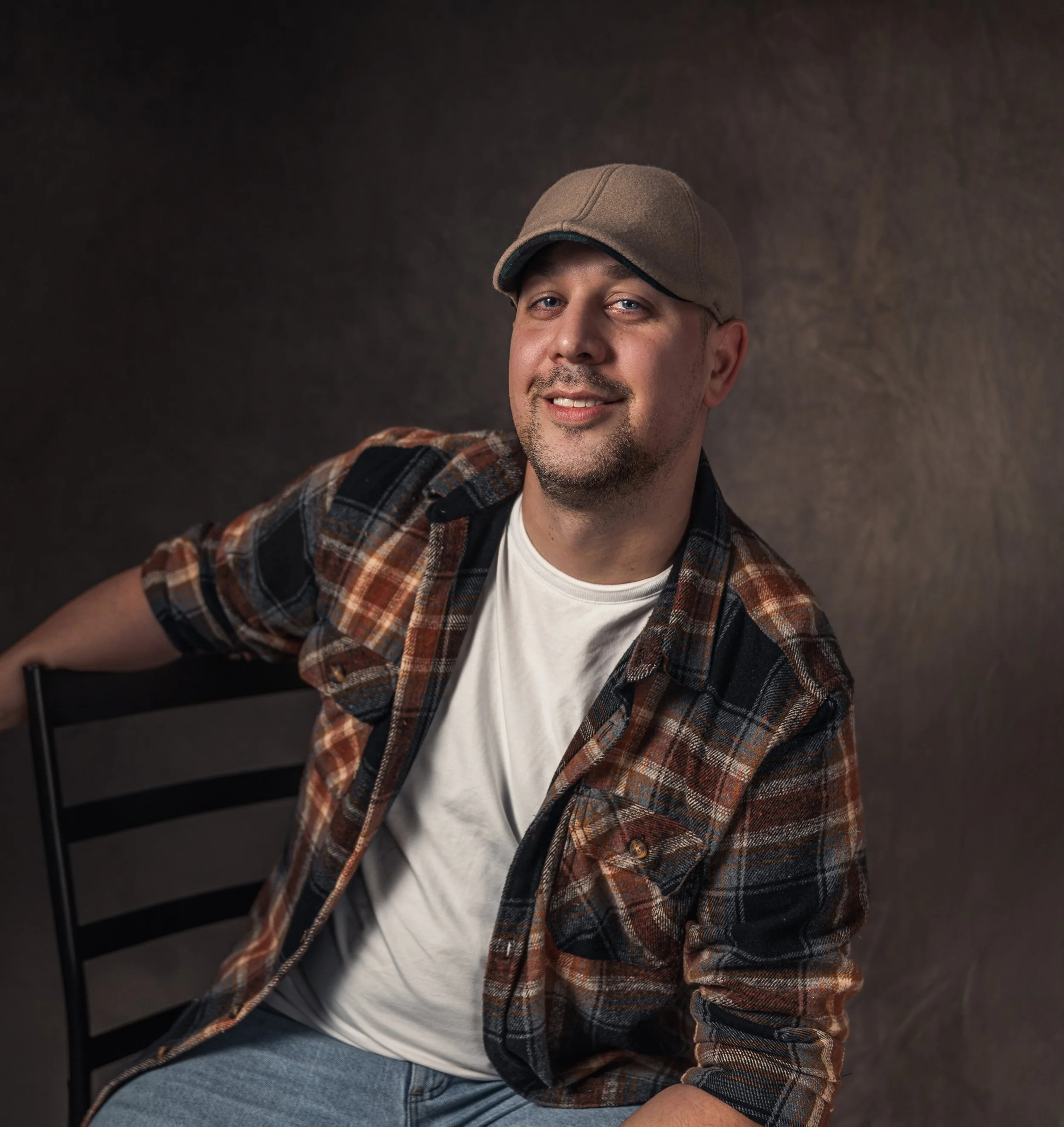 A man wearing a green cap, plaid shirt, and white t-shirt, sitting and smiling against a dark background.