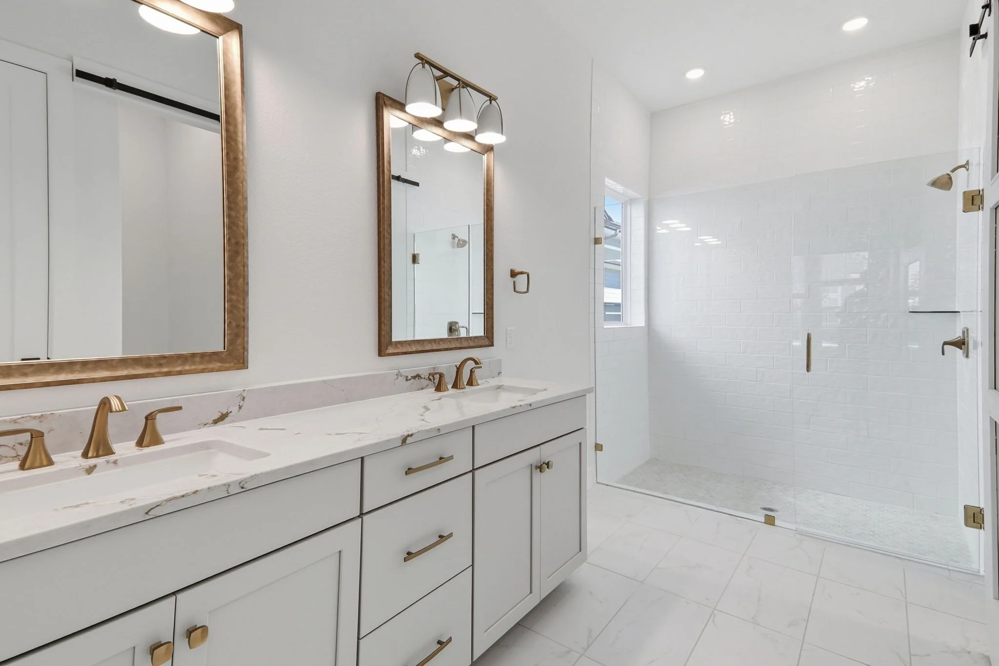 Modern bathroom with white vanity, gold fixtures, large mirrors, bright lighting, glass shower enclosure, and white tiled walls and floor.