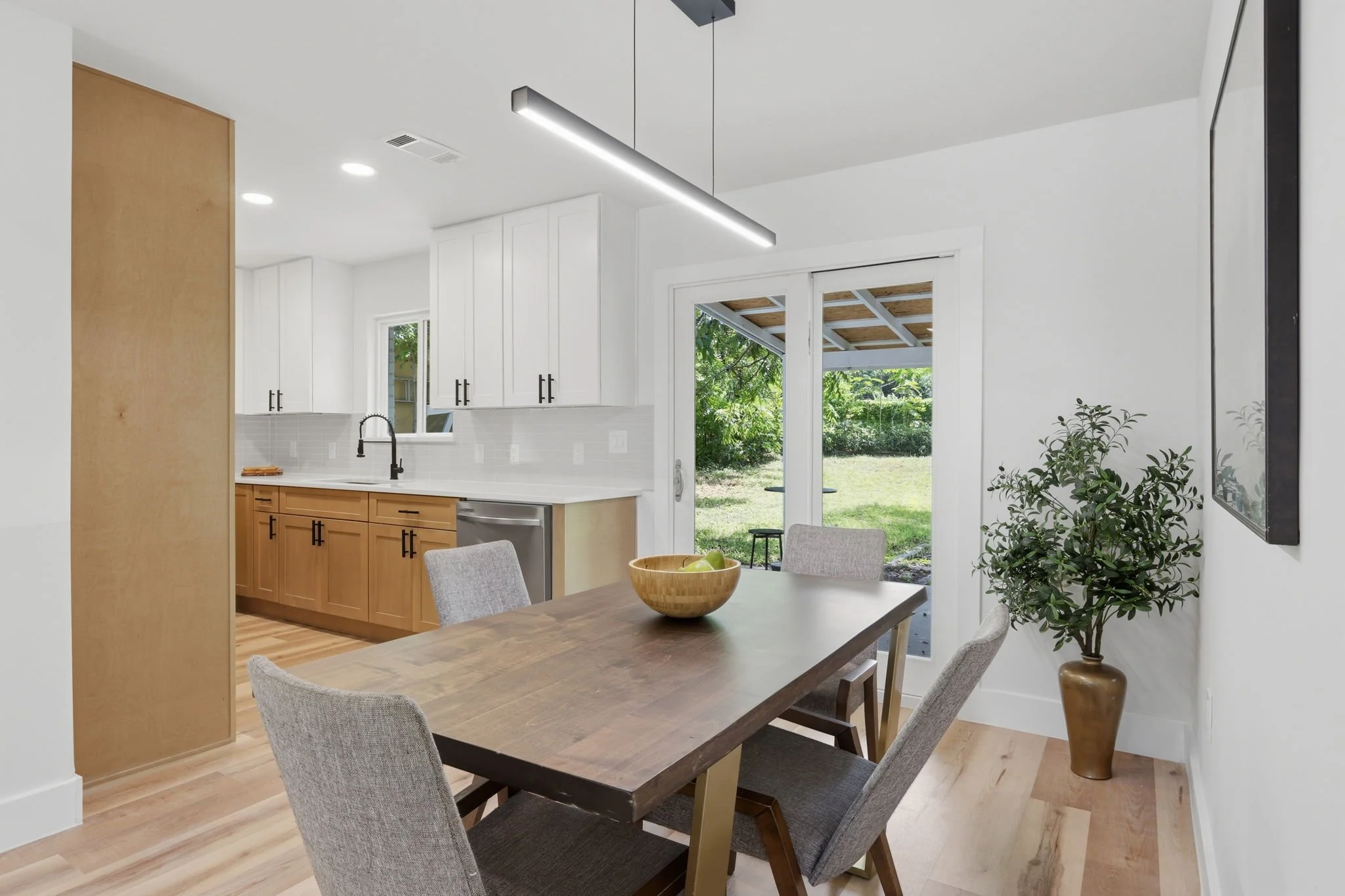 Kitchen with white upper cabinets and wood lower cabinets, black faucet at sink, sliding glass door leading outside to a backyard, and a dining table with four gray upholstered chairs and a wooden bowl with green apples.